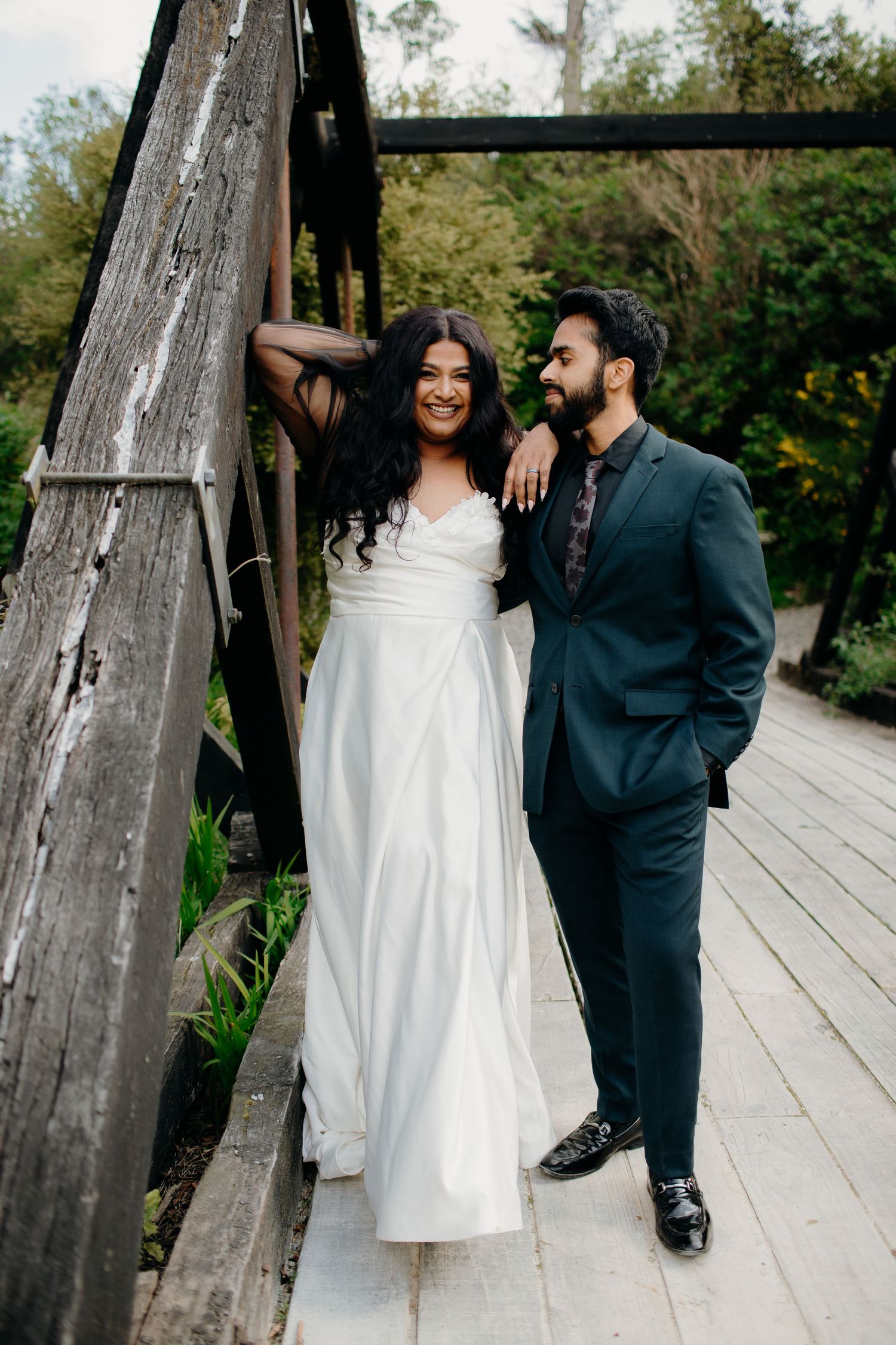 A couple poses together on a wooden boardwalk, with the bride wearing a flowing white dress and groom in navy suit.