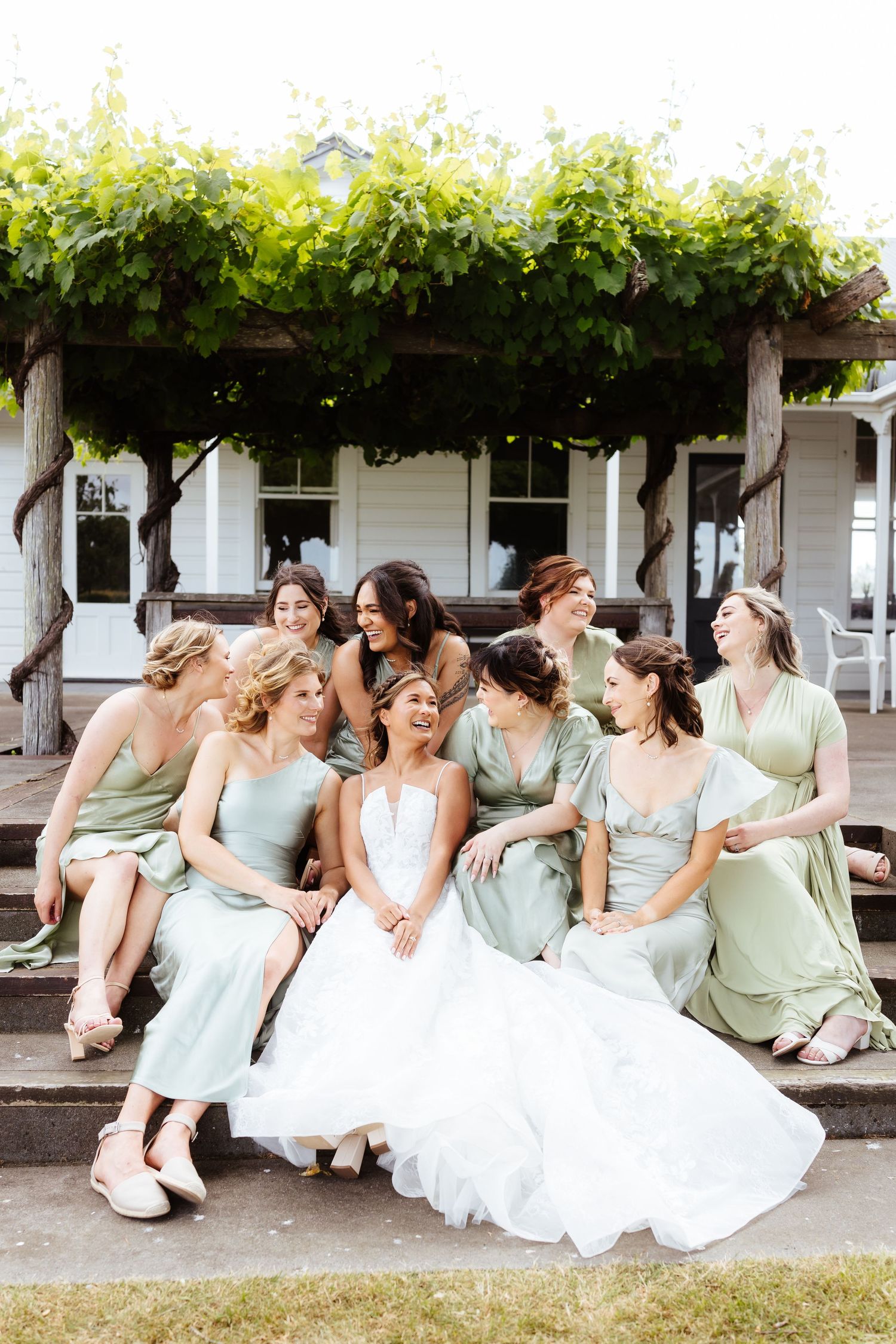 Wedding party in sage green dresses and white gown sitting and laughing together in a candid moment.