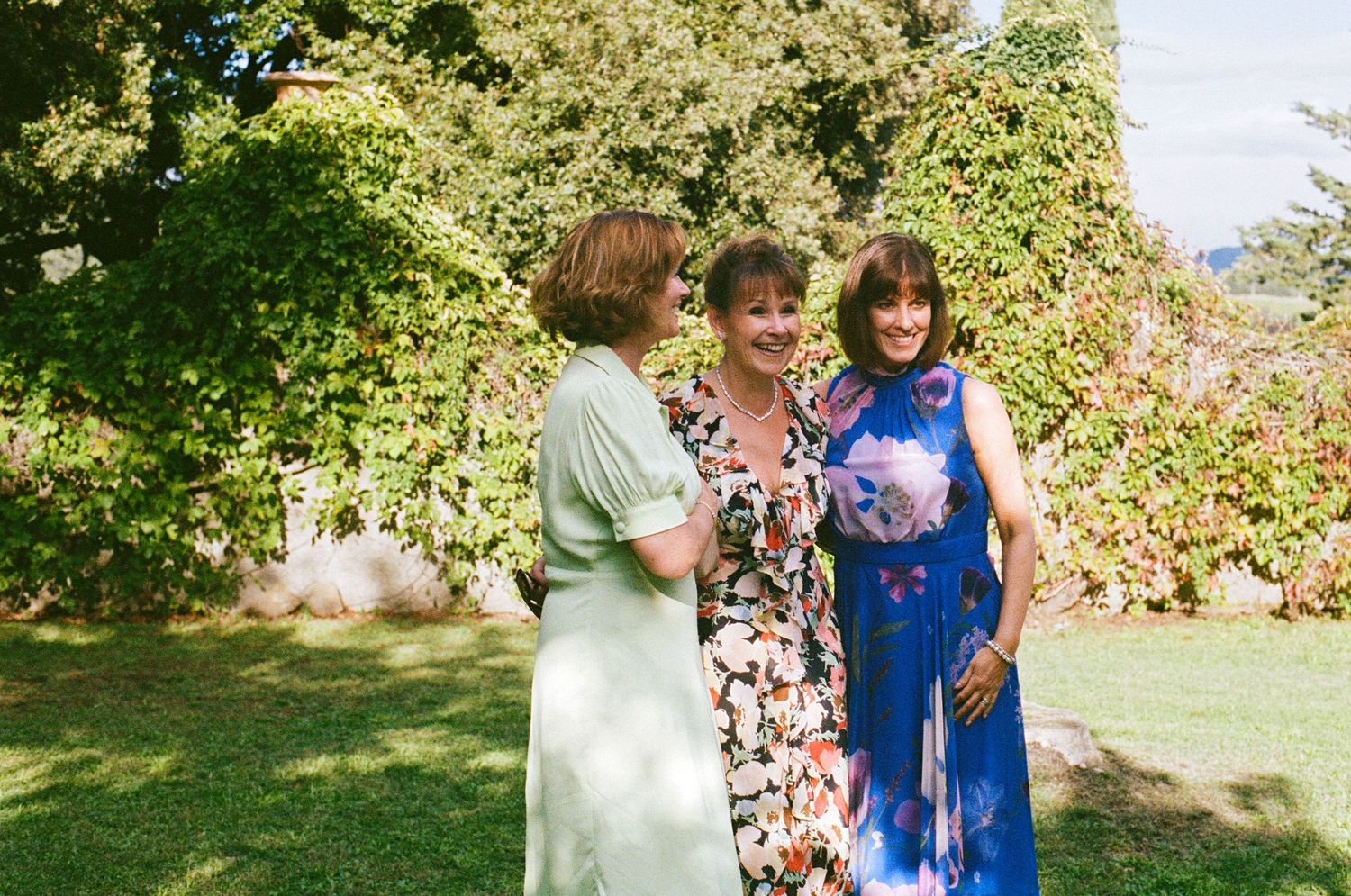 Three women smiling together in a sunlit garden, wearing elegant floral and pastel dresses at a wedding.