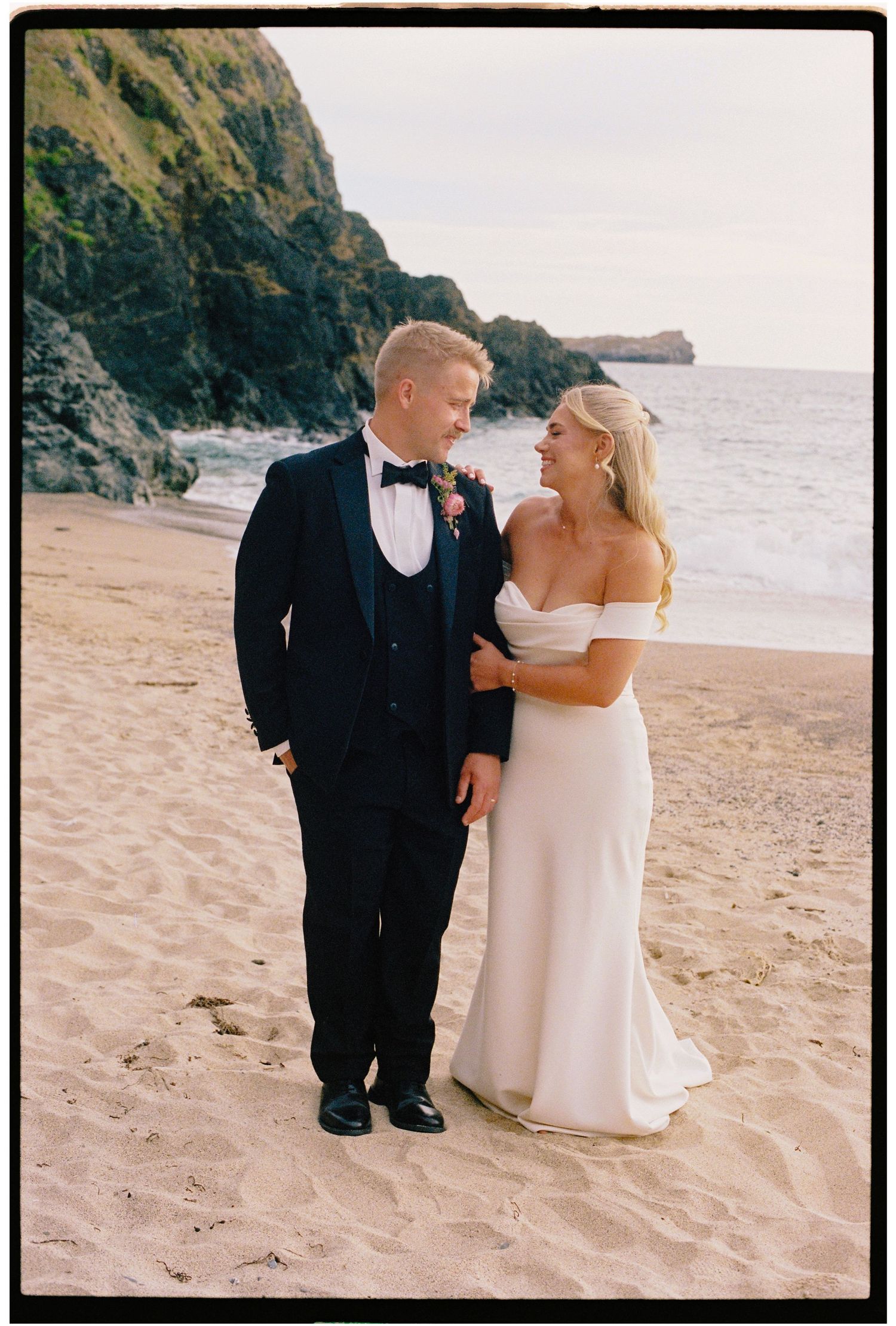 Bride and groom share a romantic moment on a sandy beach with rocky cliffs and ocean waves in the background.