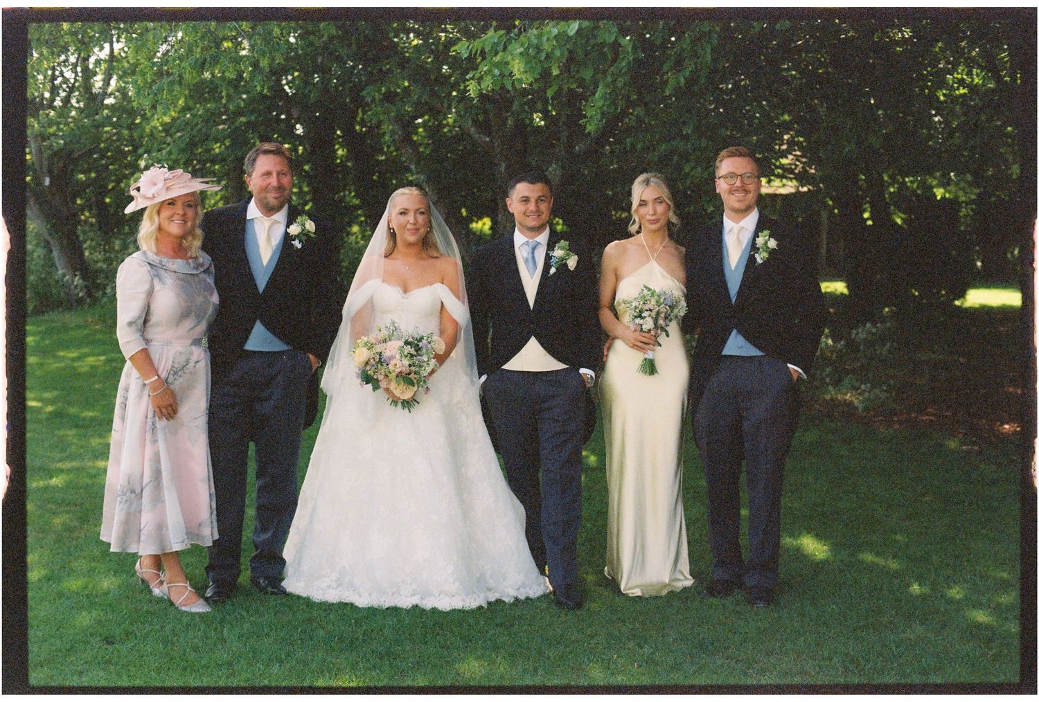 Wedding party of six posing outdoors on green lawn, bride in white gown with veil, groom in morning suit, bridesmaids and groomsmen.