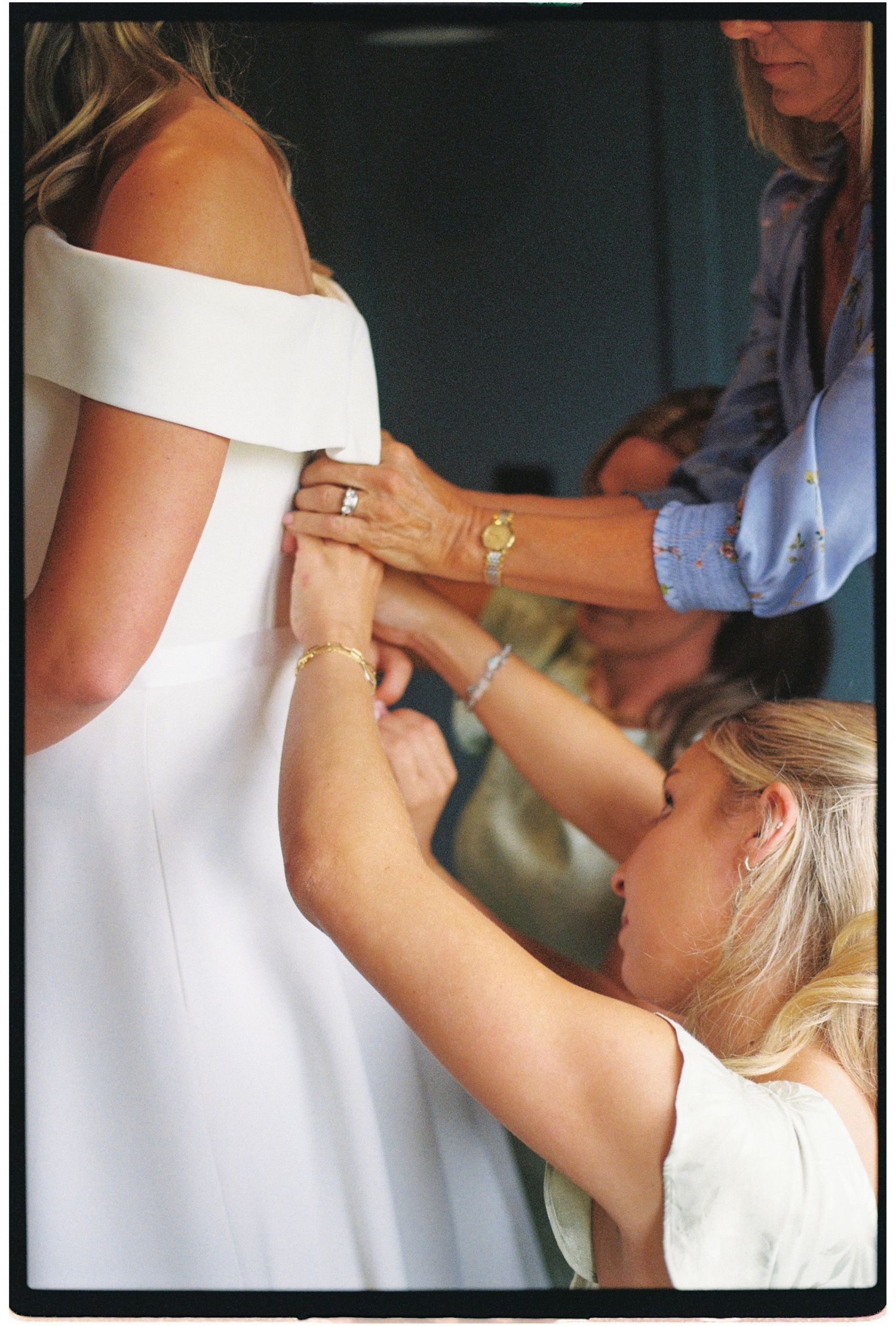 Two women helping a bride button up her white off-shoulder wedding dress before the ceremony.