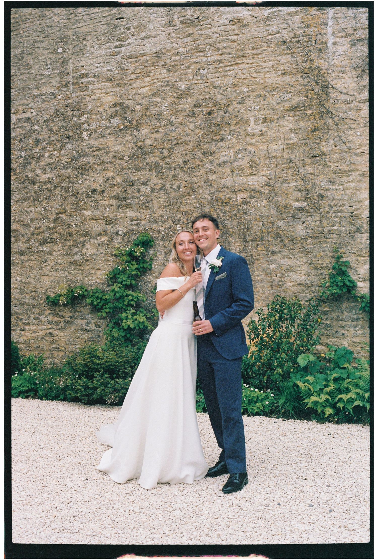 Bride in white off-shoulder gown and groom in navy suit pose together before a stone wall at an outdoor wedding.