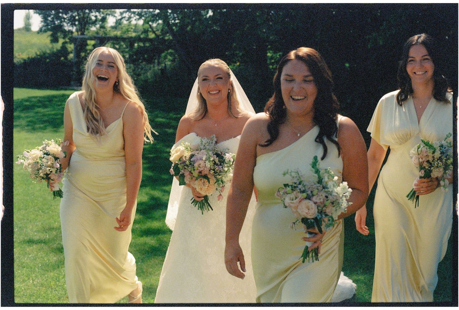 Bride in white gown walks with three bridesmaids in yellow dresses, all holding bouquets on a sunny lawn.