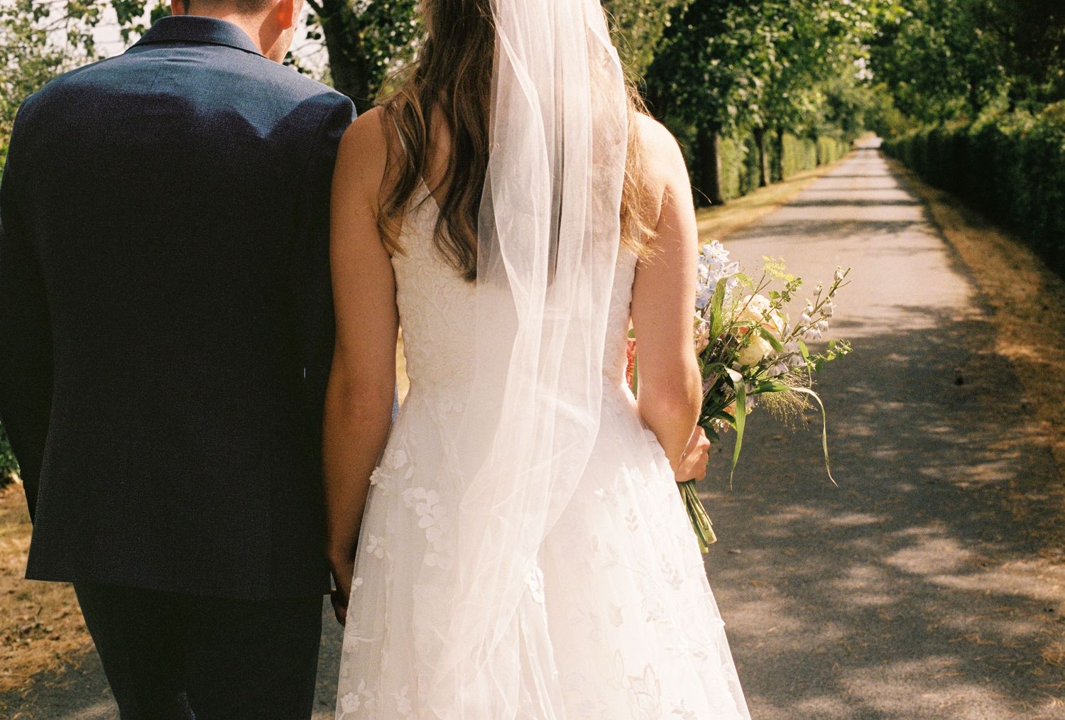 Bride and groom walking hand in hand down a tree-lined path, bride wearing white gown with veil and bouquet.