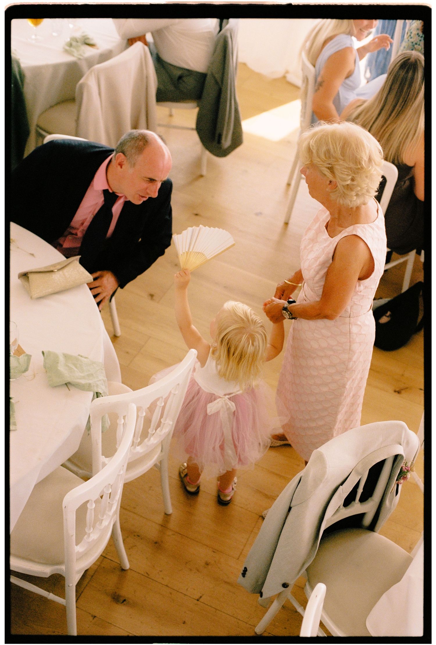A young girl in a pink tutu waves a fan at a wedding reception while an elderly couple looks on warmly.