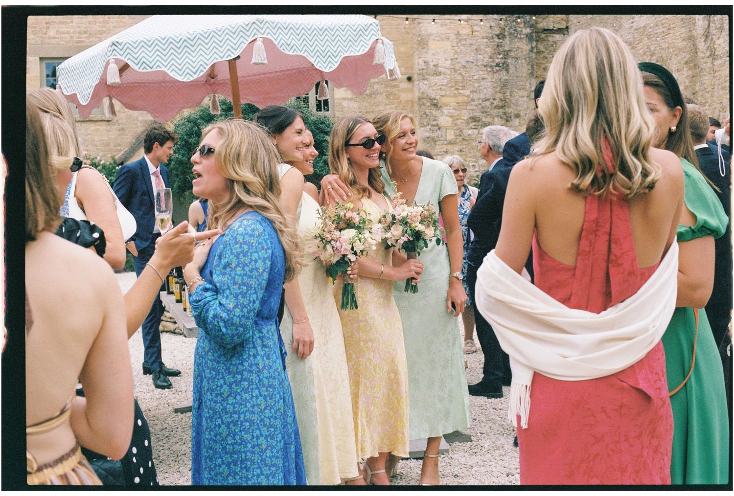 Guests in colorful summer dresses gather outdoors at a wedding celebration, with a woman holding a floral bouquet.