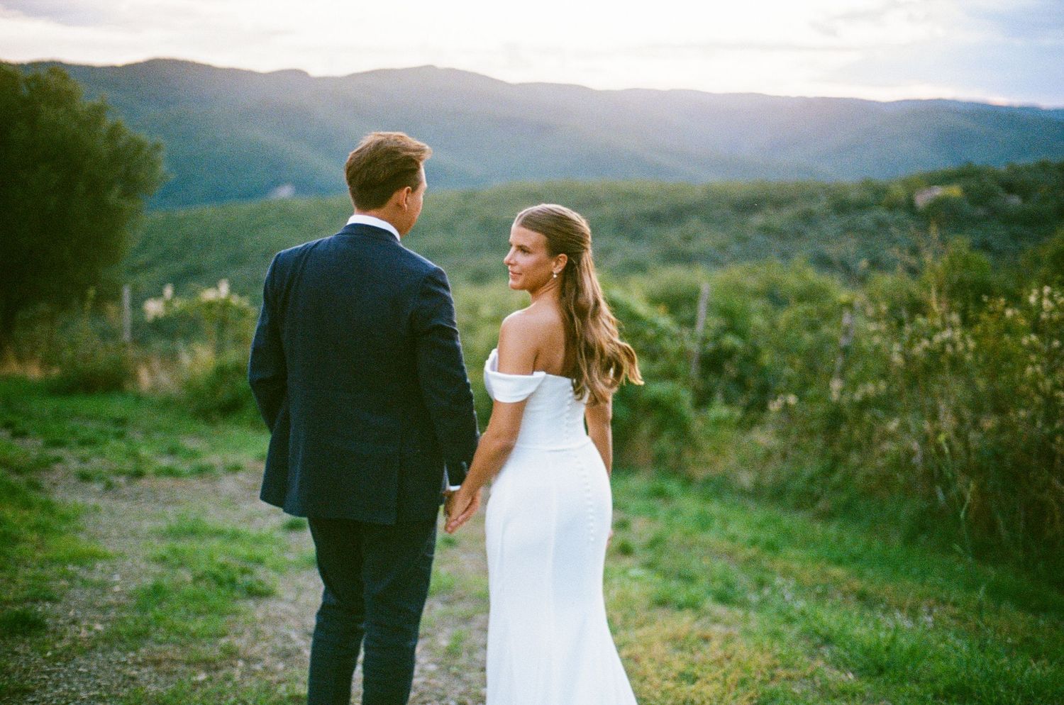 Bride in off-shoulder gown and groom in navy suit holding hands overlooking scenic Tuscan mountain landscape.