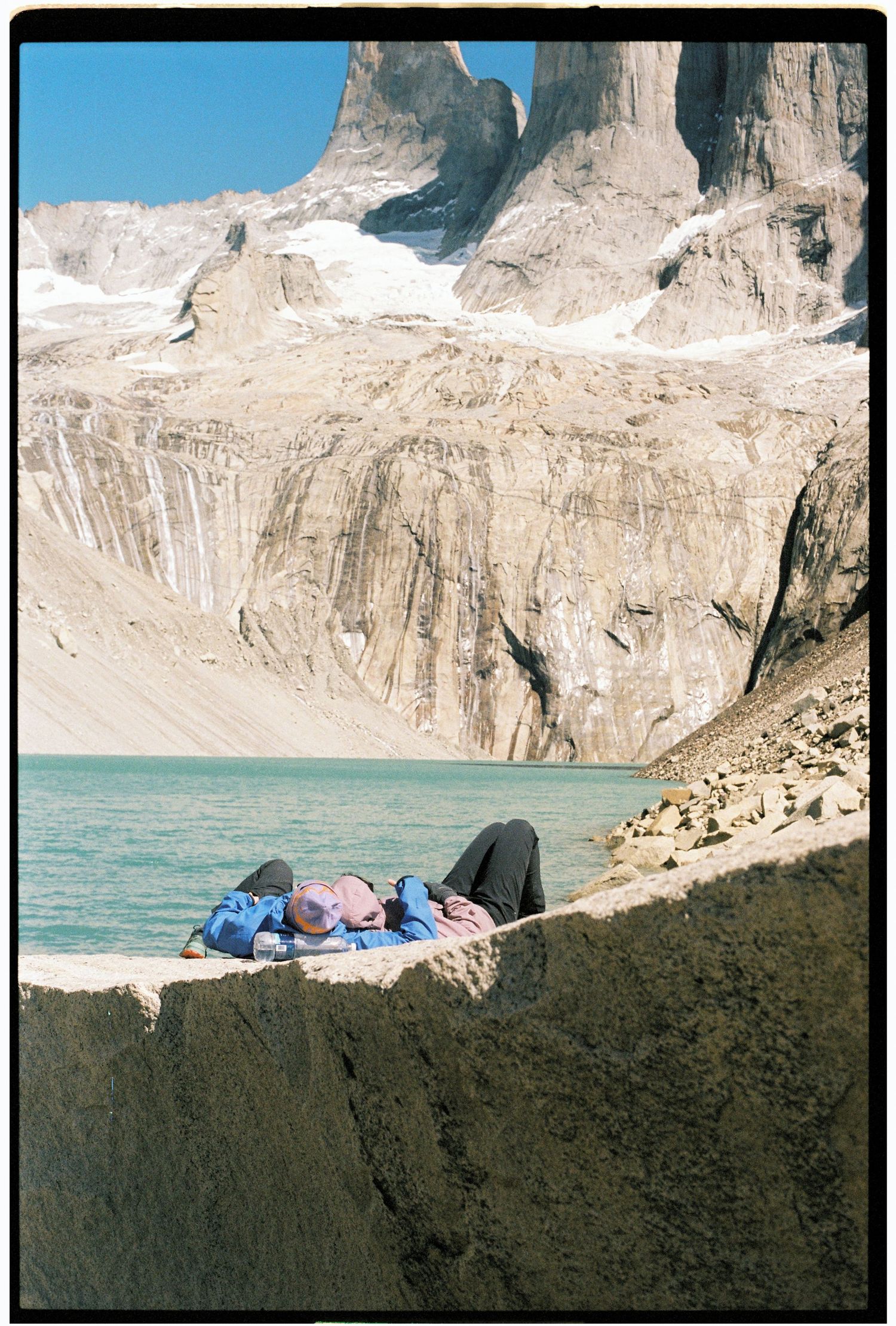 Two people resting on rocky ledge beside turquoise glacial lake below towering Patagonia mountain peaks.