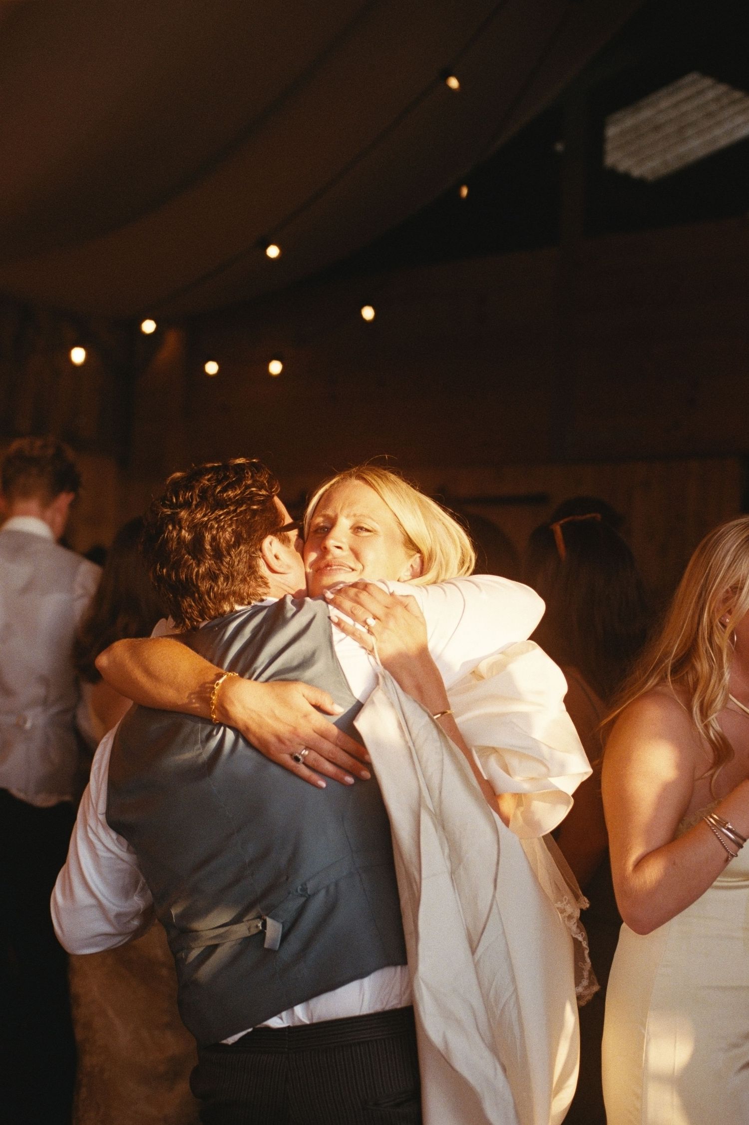Bride and groom share a joyful embrace on the dance floor at their wedding reception under warm string lights.