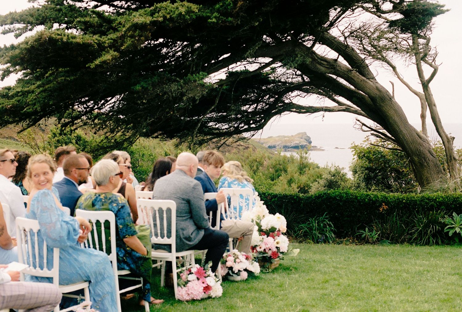 Outdoor wedding ceremony with guests seated on white chairs under a large tree overlooking the ocean.