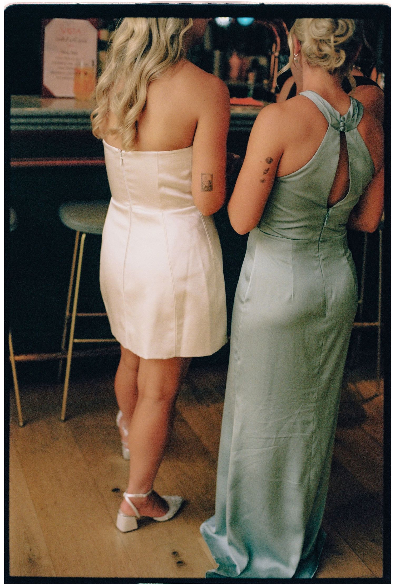 Two women in elegant dresses stand at a bar, one in white mini dress, other in sage green gown, both with tattoos.