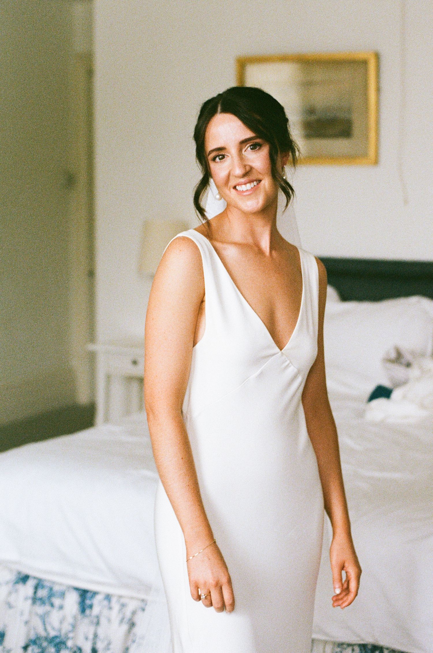 Smiling bride in elegant sleeveless white V-neck wedding gown standing in a bright bedroom.