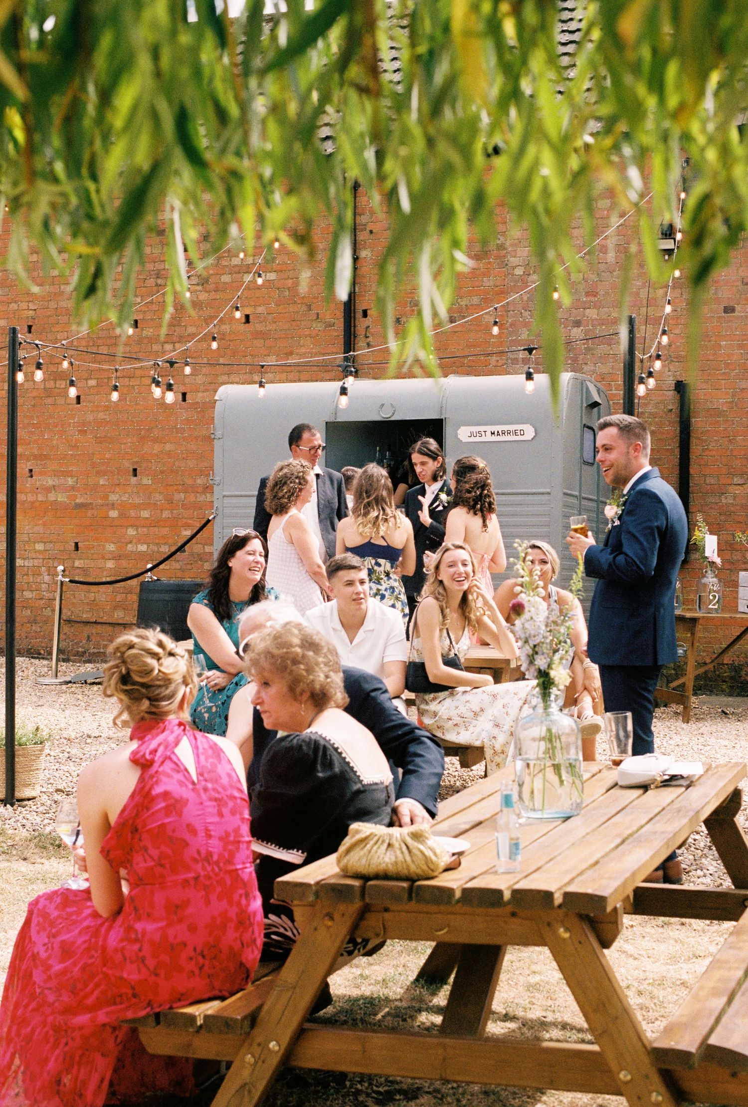 Outdoor wedding reception with guests at picnic tables, string lights, and a brick wall backdrop on a sunny day.