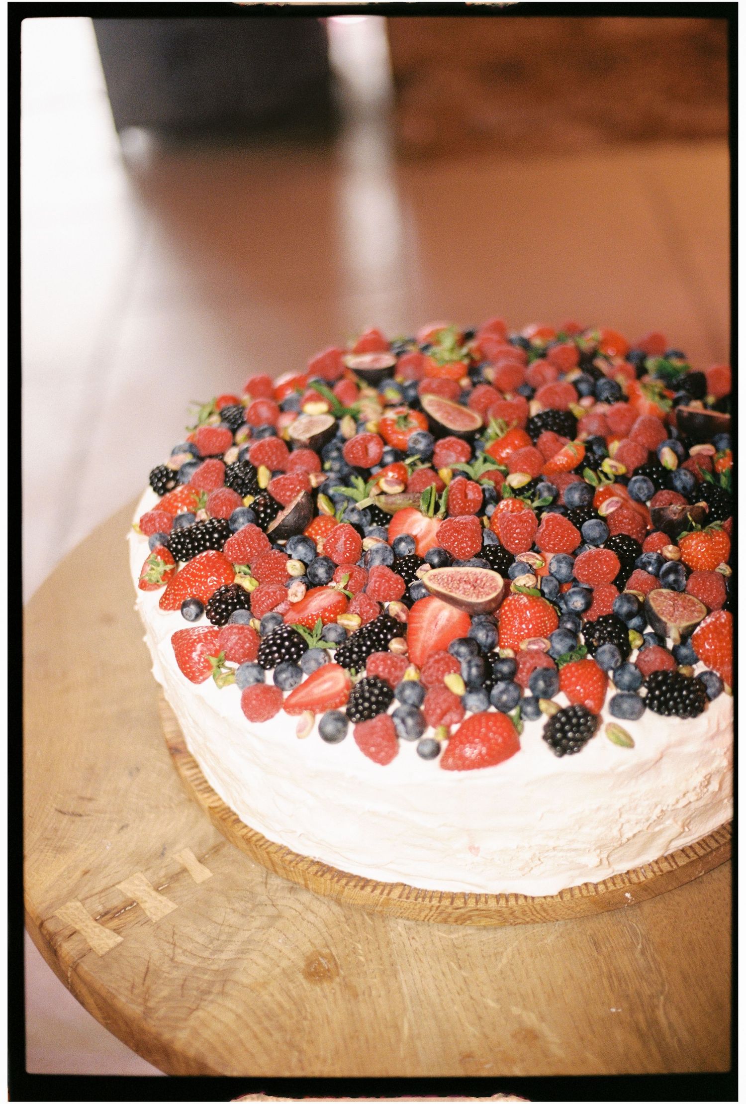 White frosted cake topped with fresh strawberries, blueberries, blackberries, and figs on a wooden cake stand.
