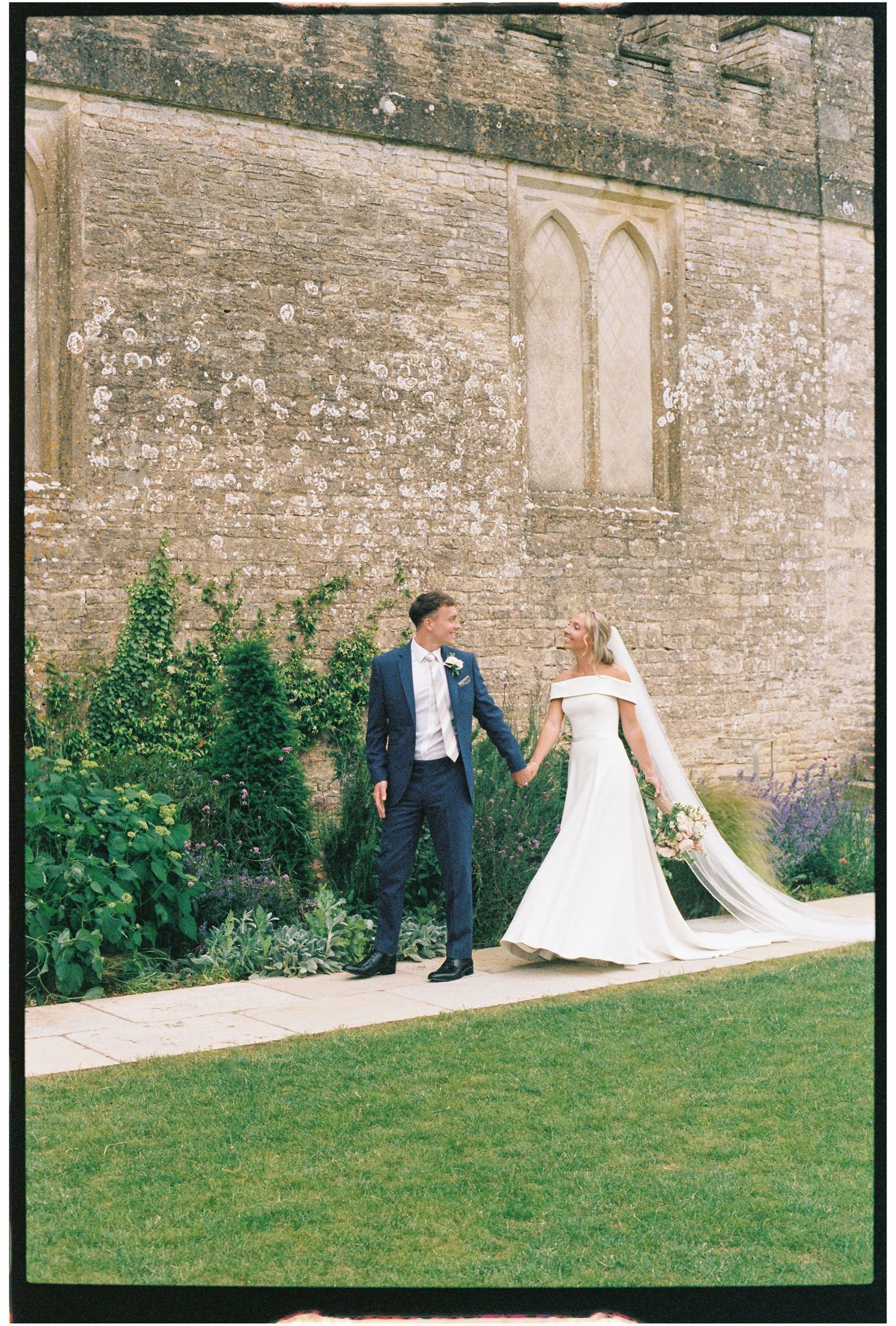 Bride and groom holding hands walking beside a historic stone church wall with lush green gardens on their wedding day.