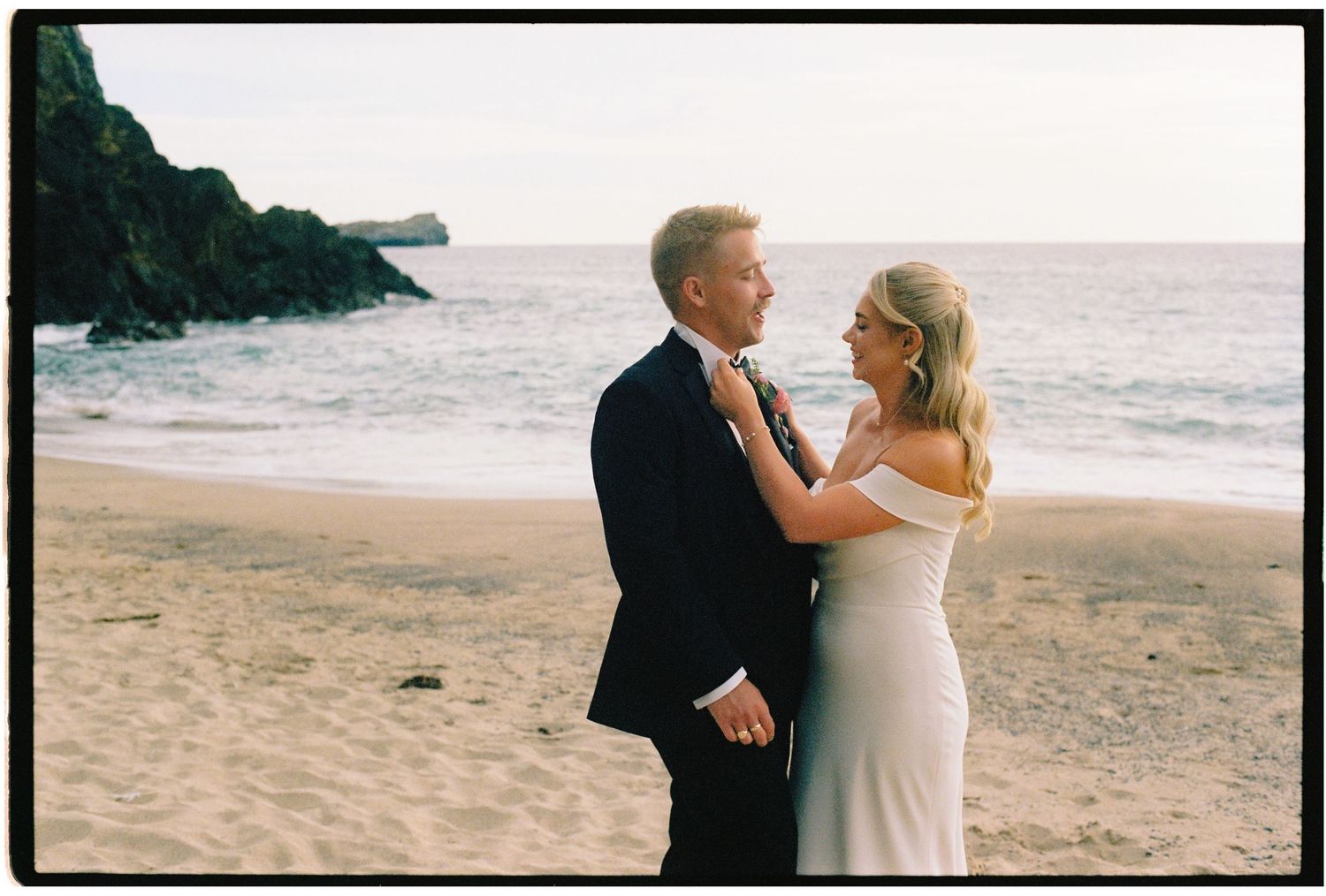 Couple in wedding attire sharing a romantic moment on a sandy beach with ocean waves and rocky cliffs in the background.