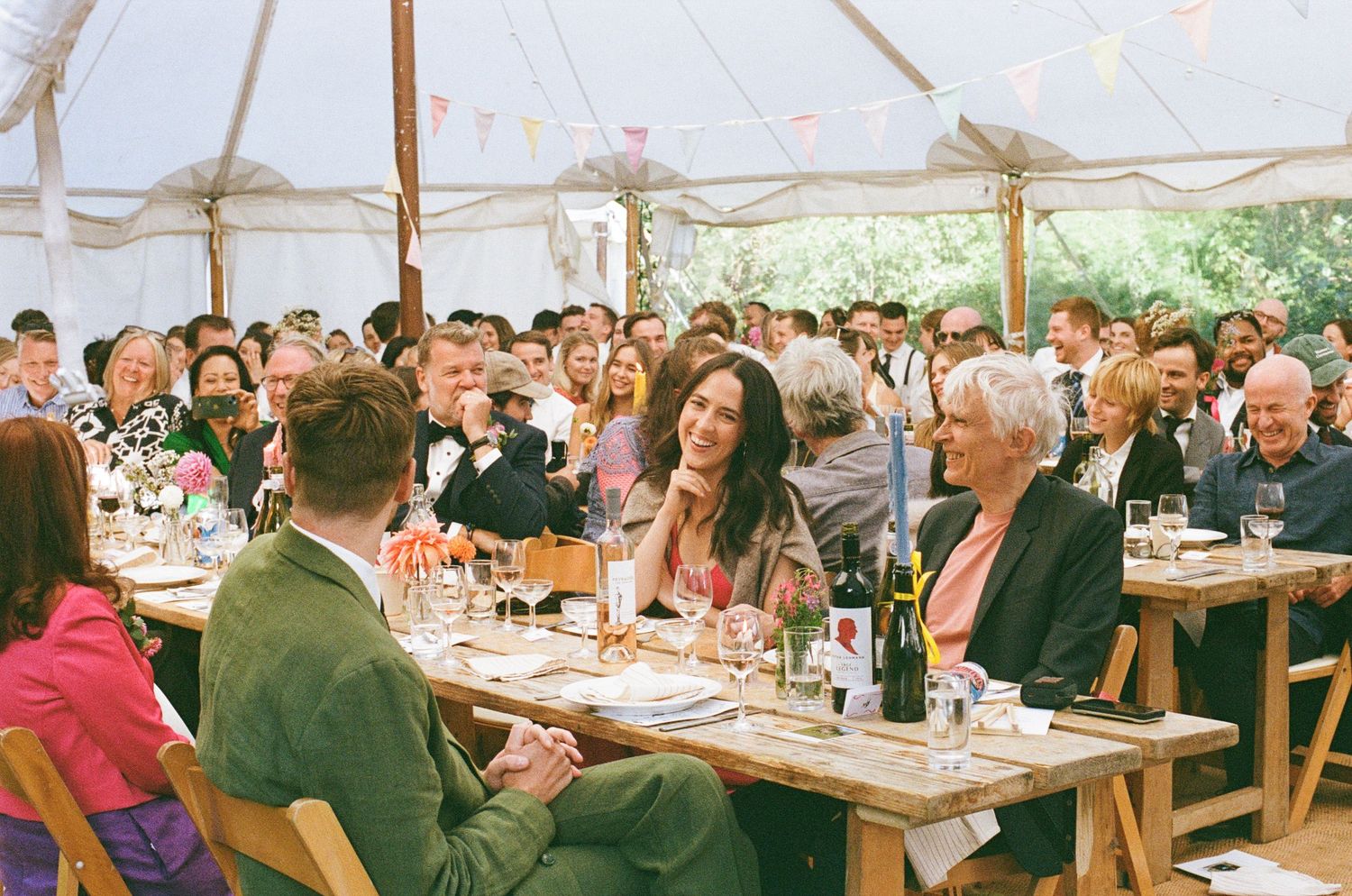 Wedding reception guests laughing and socializing at long wooden tables inside a decorated marquee tent.