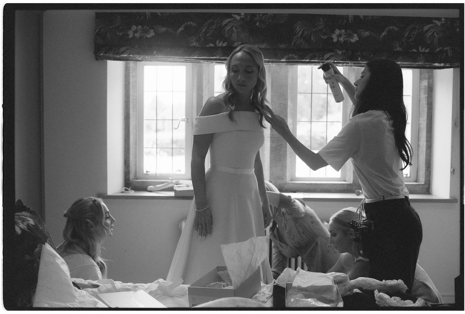 Black and white photo of a bride being helped into her wedding dress by bridesmaids in a sunlit room.