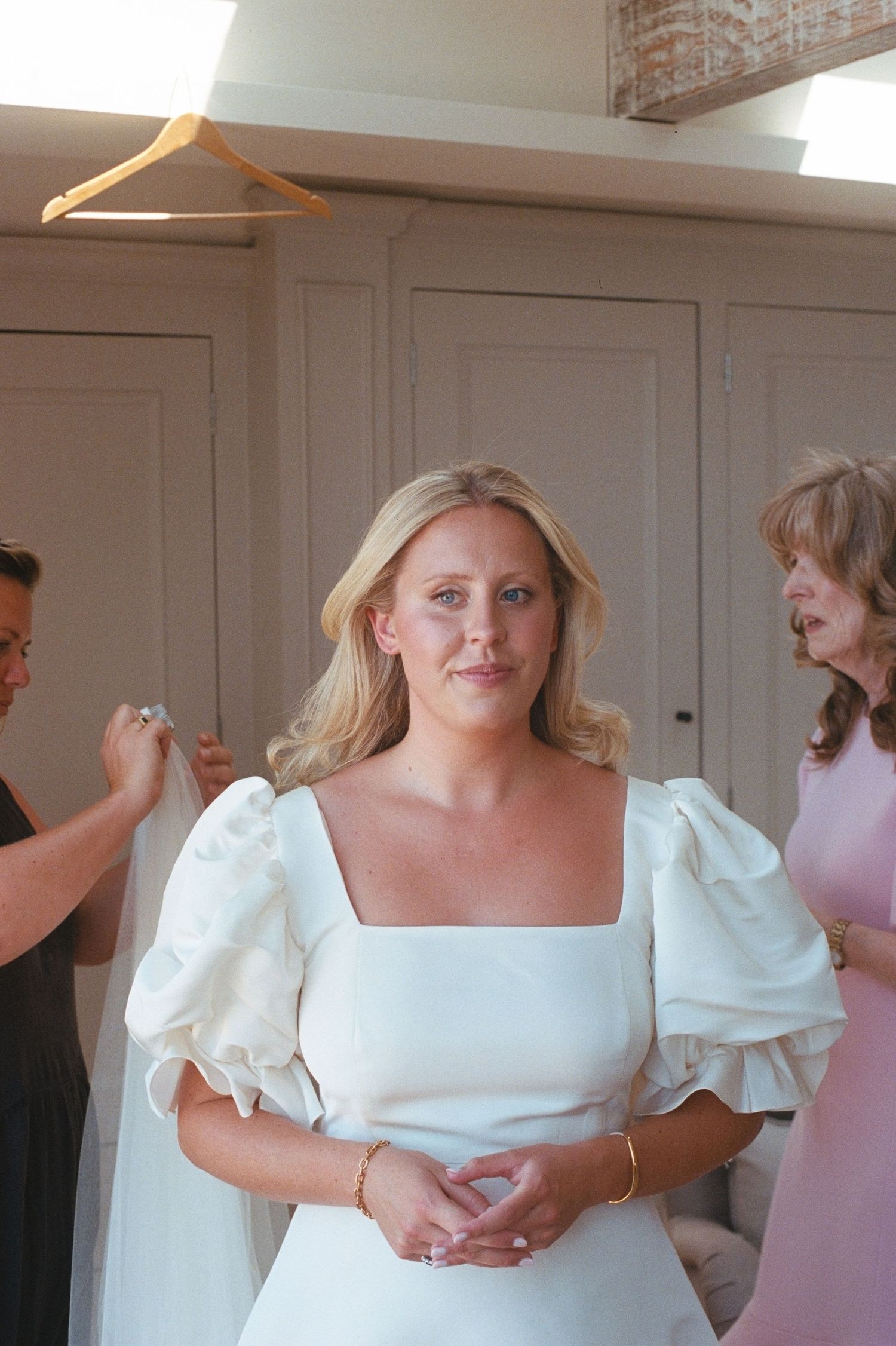 Blonde bride in white puff sleeve wedding dress being helped by attendants in a bright dressing room.
