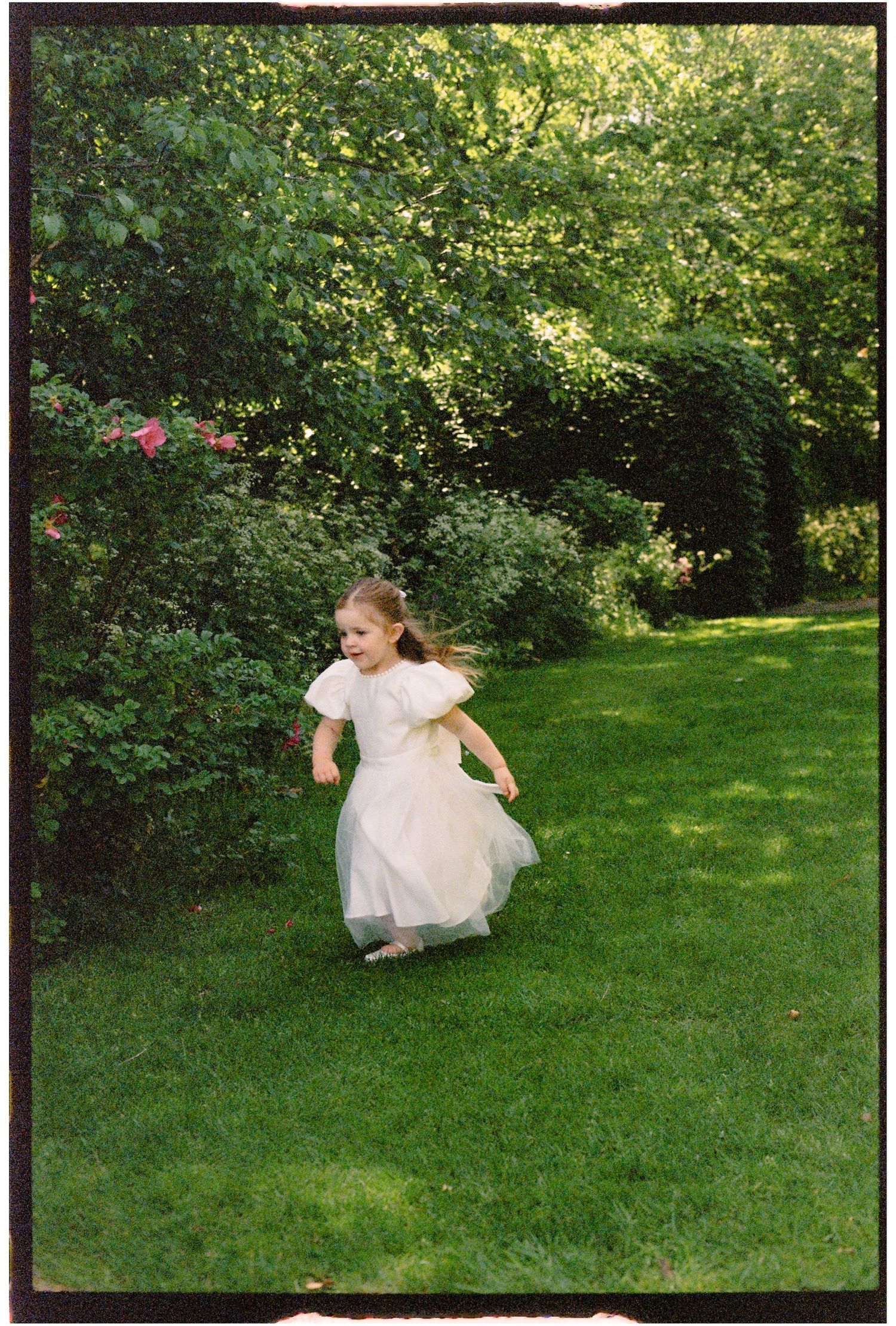 A young girl in a white dress runs joyfully across a lush green lawn surrounded by leafy garden shrubs.