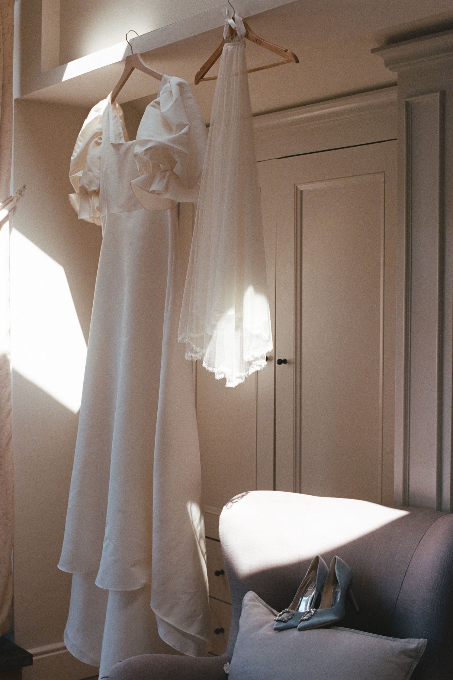 White wedding dress and veil hanging on wooden hangers near a sunlit window with heels on a chair below.