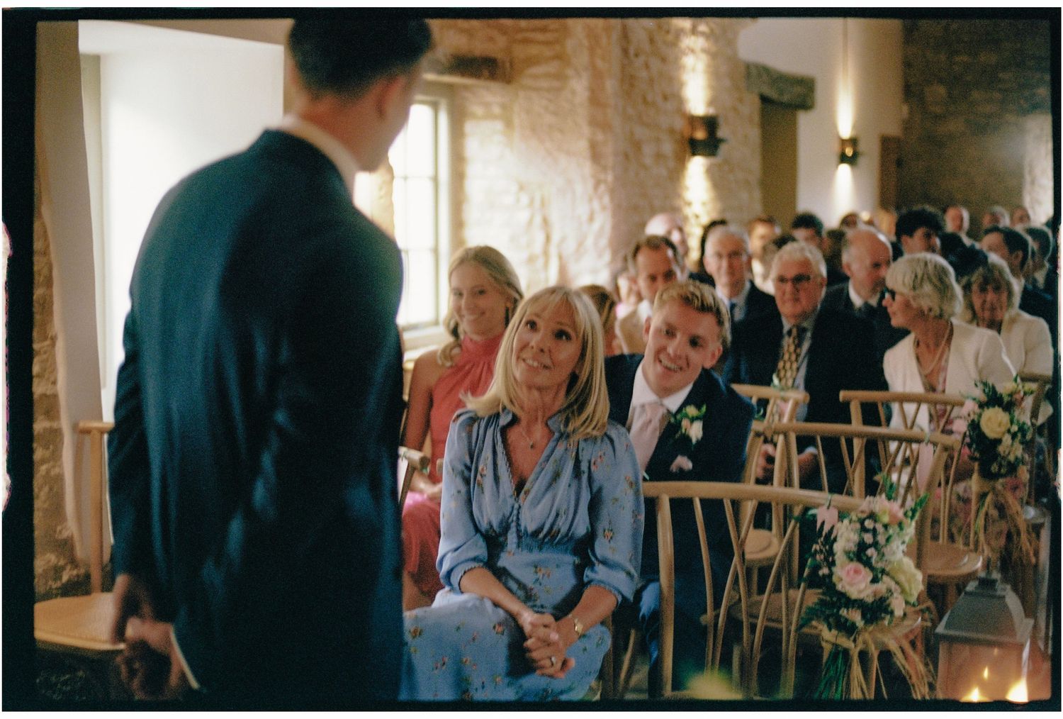 Guests smile warmly at a wedding ceremony inside a rustic stone venue with wooden chairs and floral decorations.