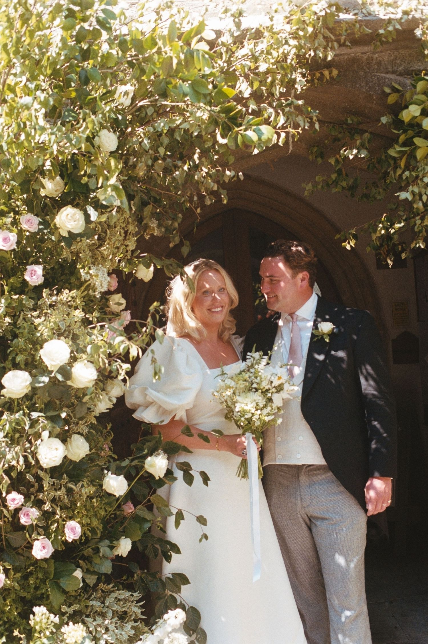 Bride and groom smiling together under a rose-covered arch, bride holding a white bouquet in a puff-sleeve wedding gown.