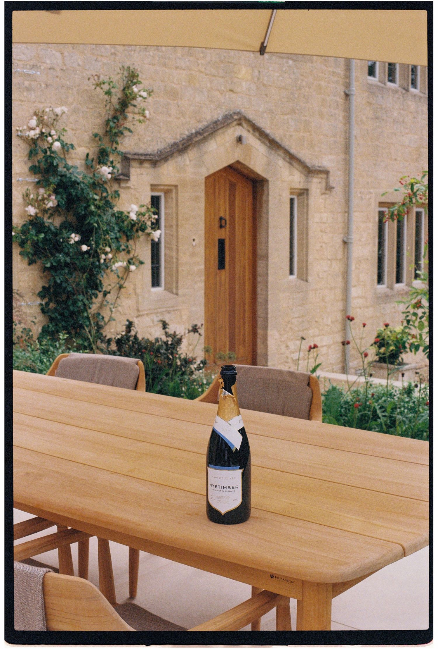 A bottle of champagne on a wooden outdoor dining table beside a charming stone cottage with a timber door.