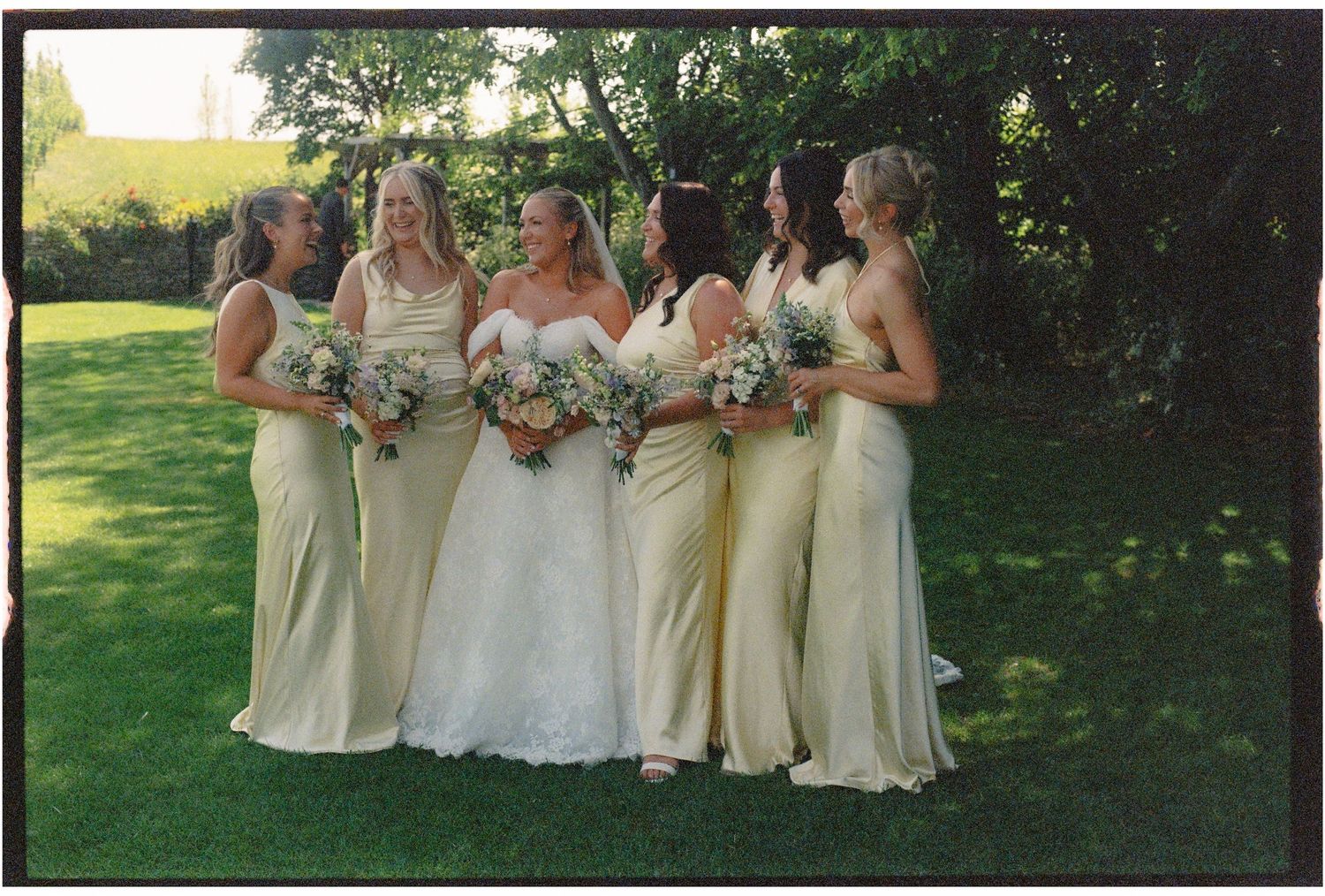 Bride in white lace gown with four bridesmaids in yellow dresses holding bouquets outdoors on green lawn.