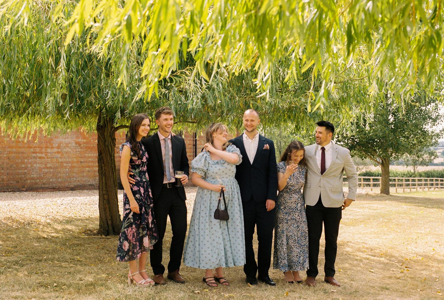 Family of six posing outdoors under a willow tree on a sunny day, dressed in formal attire for a special occasion.