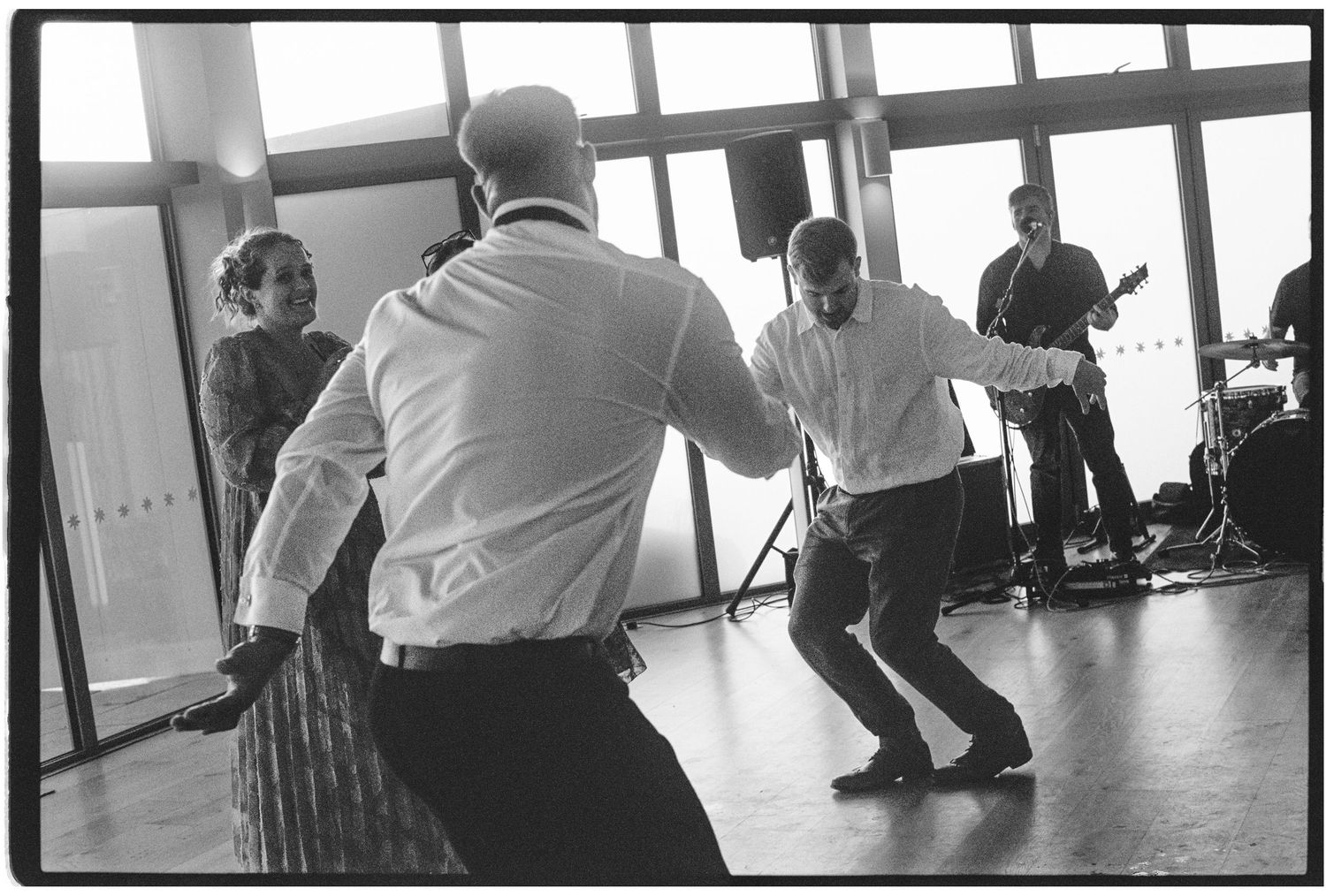 Black and white photo of men dancing energetically at a social event with a guitarist performing in the background.