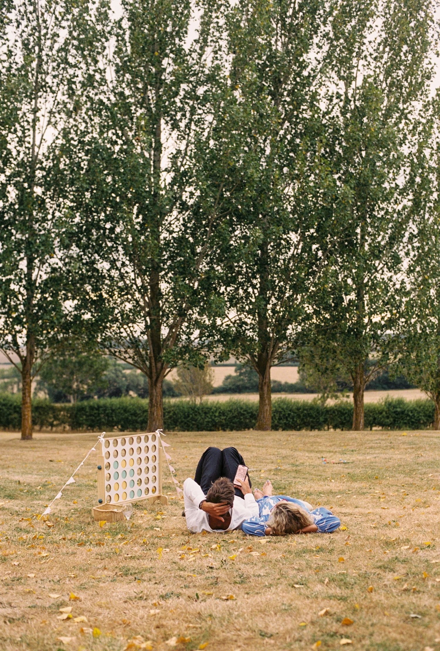 Couple relaxing on grass in a park with a giant Connect Four game and tall poplar trees in the background.