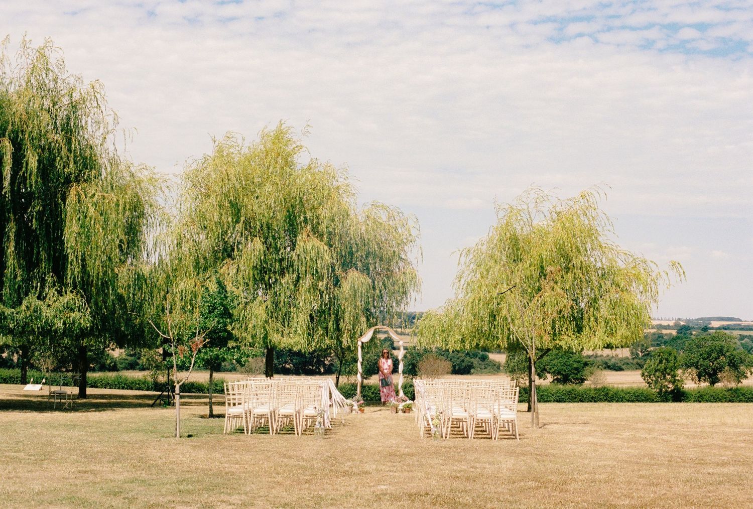 Outdoor wedding ceremony setup with white chairs and floral arch between weeping willow trees on a sunny green lawn.