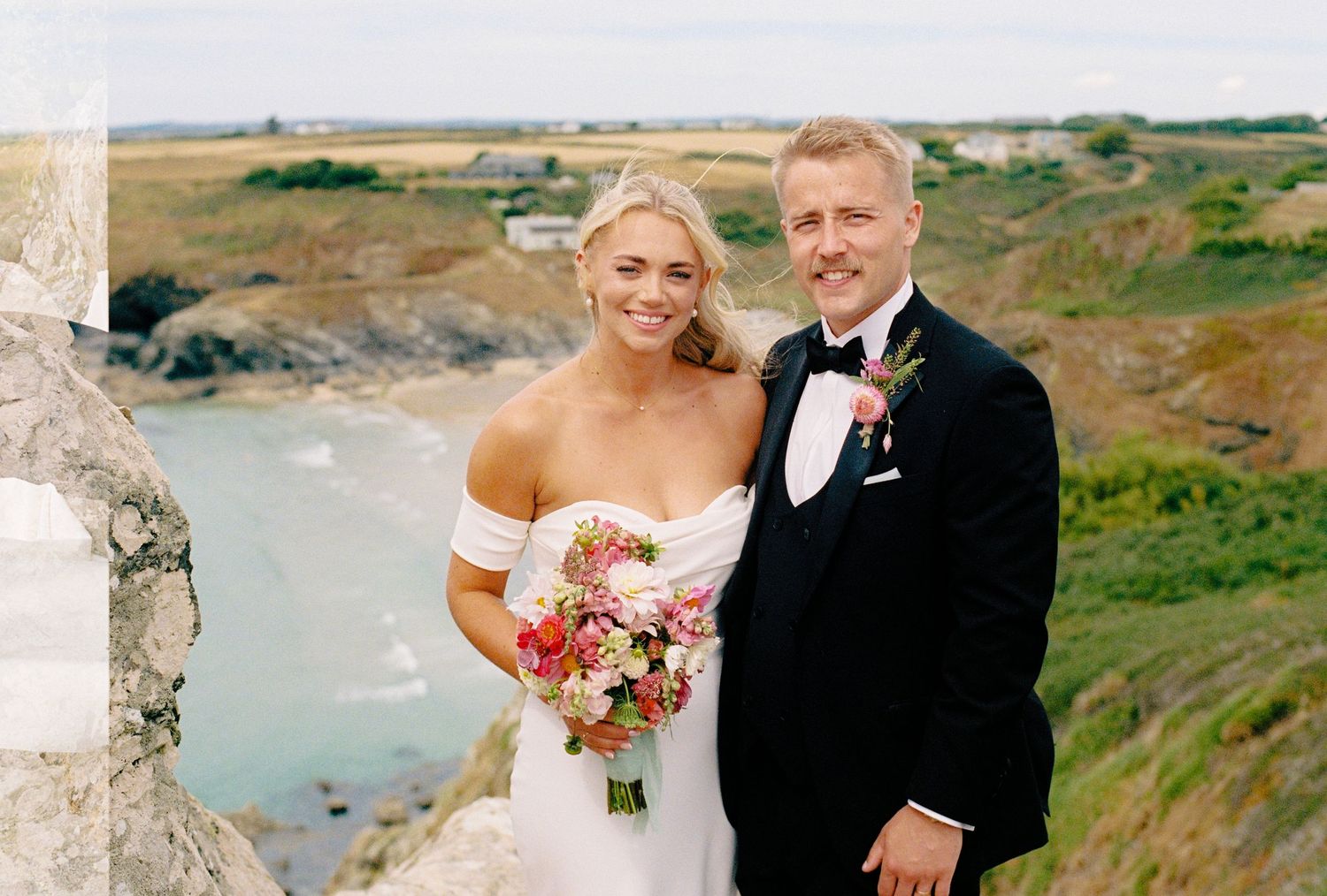Bride and groom smiling together on coastal cliffs, bride holding colorful bouquet in white off-shoulder gown.