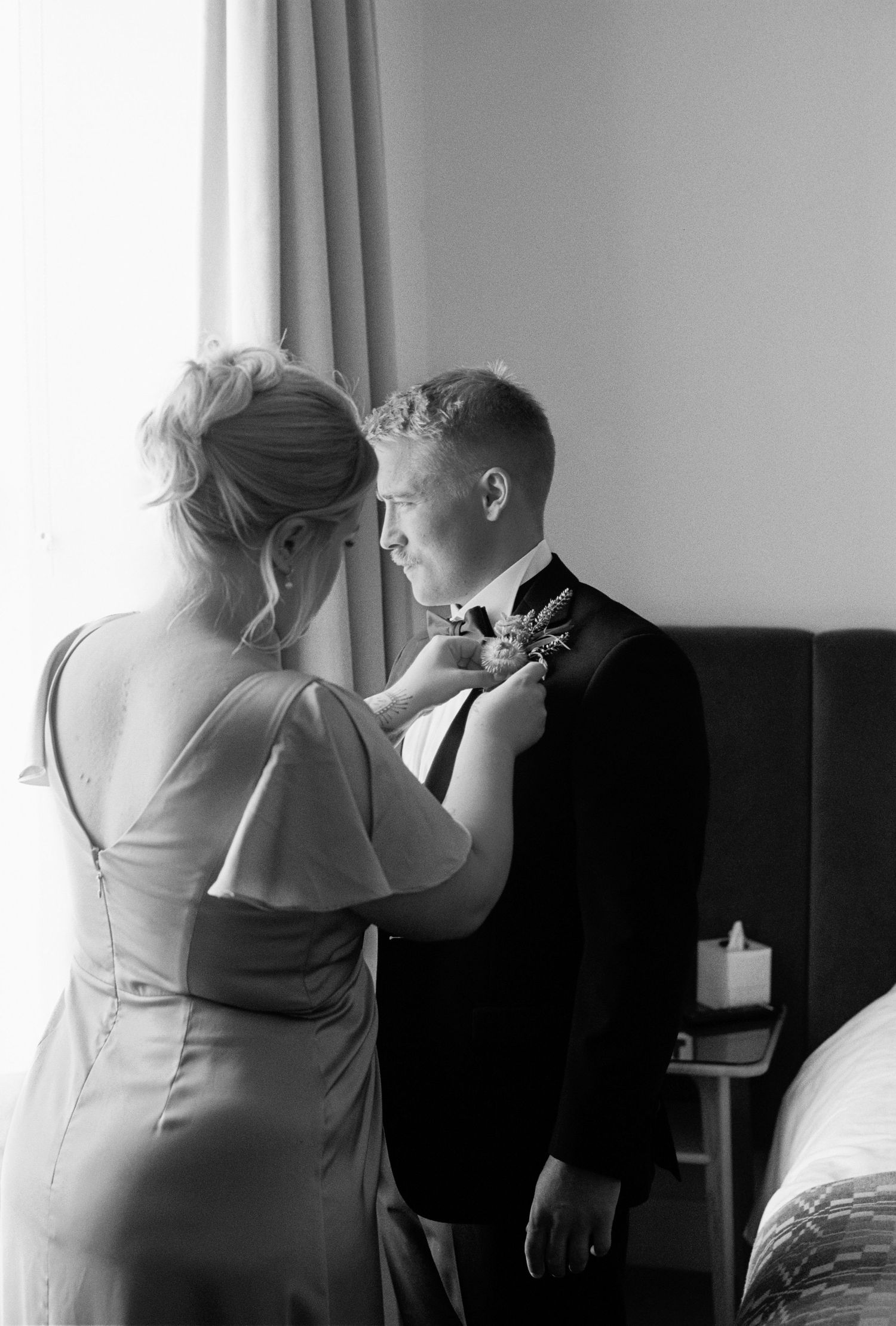 Black and white photo of a woman pinning a boutonniere on a groom in a suit before their wedding ceremony.