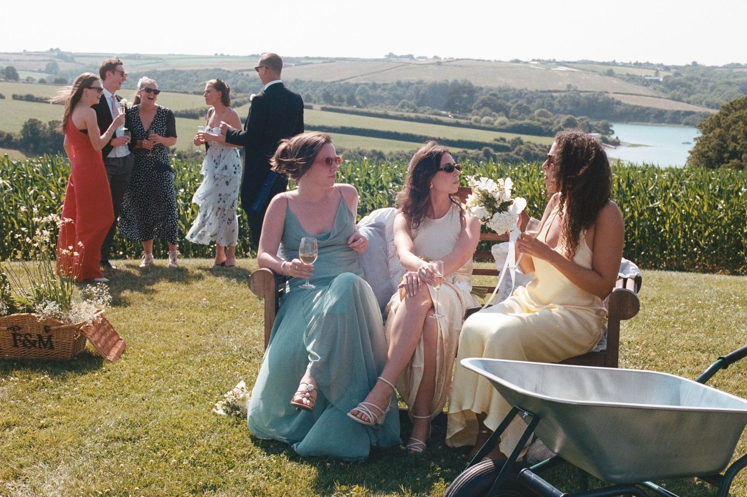 Bridesmaids in elegant gowns laughing outdoors at a sunny countryside wedding with guests and a lake in the background.