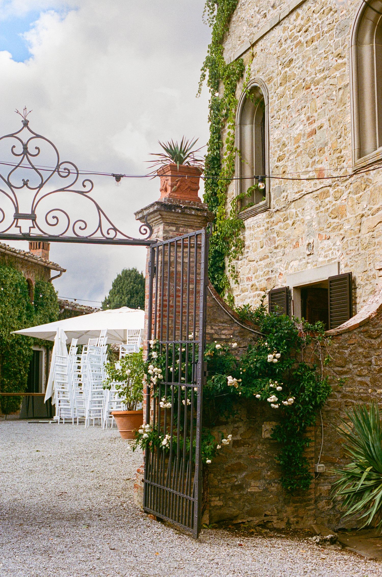 Elegant Tuscan stone villa wedding venue with iron gate, climbing ivy, and white chairs visible beyond.