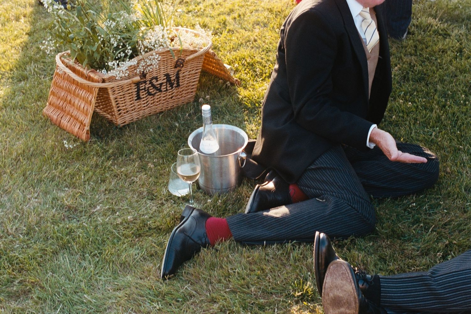 Two men in suits enjoy a Fortnum & Mason picnic on grass with champagne and wicker hamper.