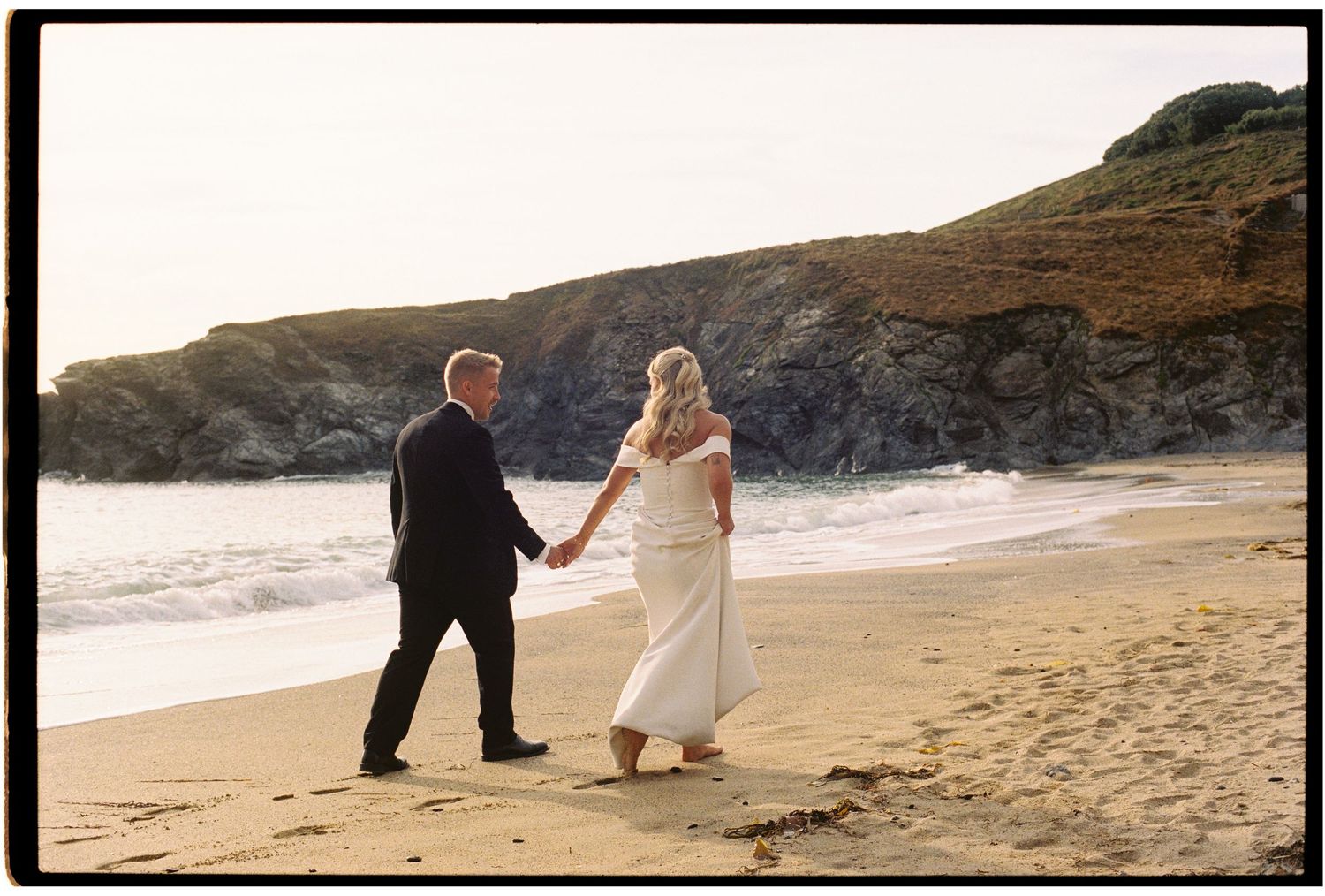 Newlywed couple holding hands walking along a scenic sandy beach with rocky cliffs at sunset.