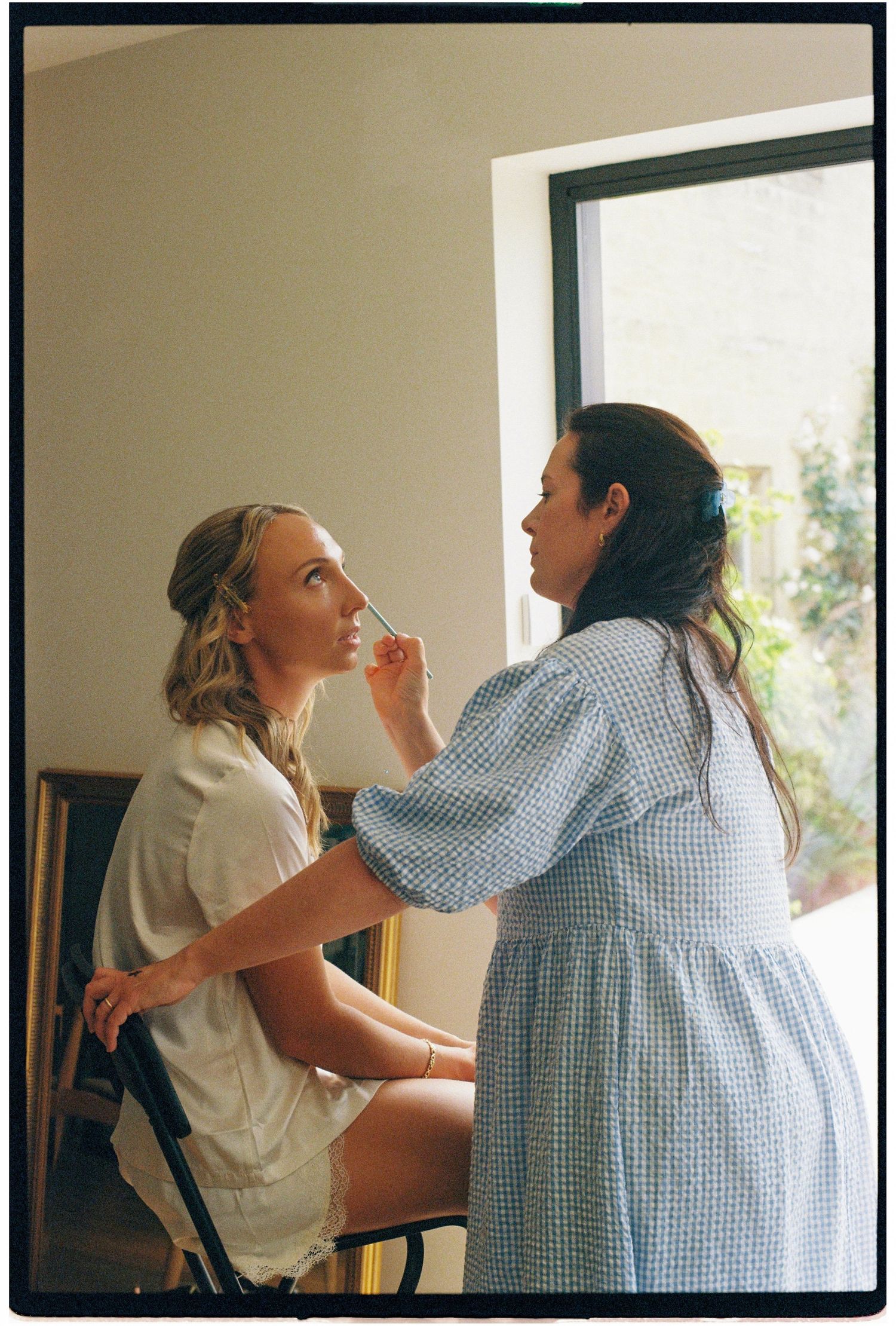Makeup artist in blue dress applying makeup to blonde woman in white outfit near bright window.