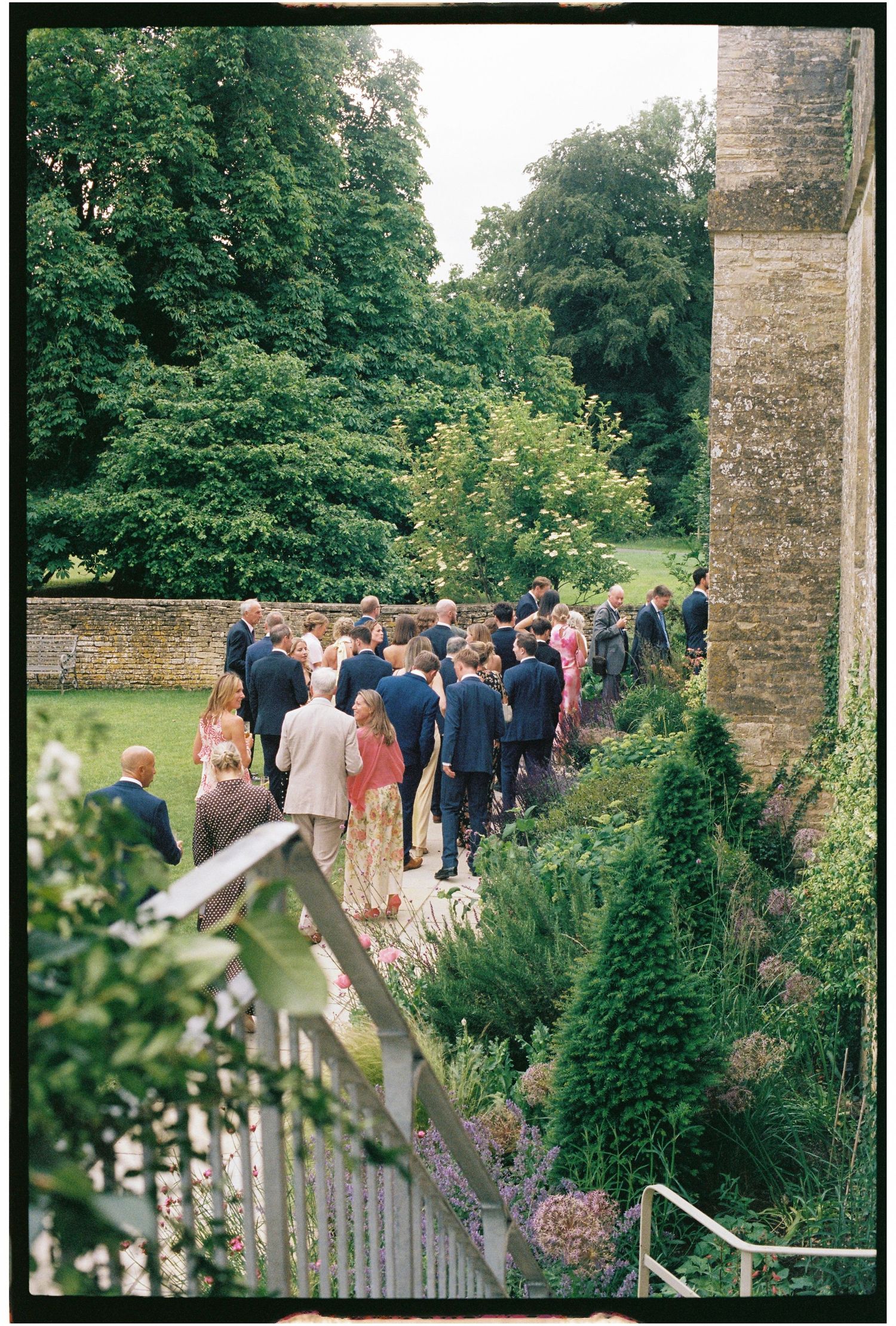 Wedding guests walk along a garden path beside a stone building, surrounded by lush greenery and flowering borders.