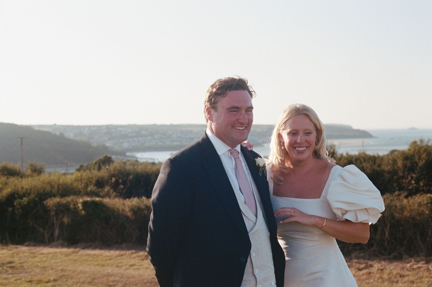 Happy couple in wedding attire smiling outdoors with coastal scenery and clear sky in the background.