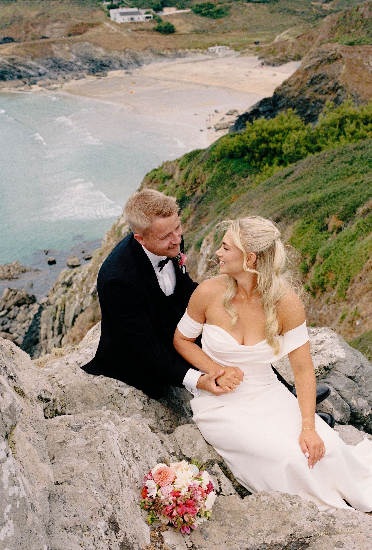 Bride and groom sitting on coastal cliffs in wedding attire with bouquet, ocean beach backdrop below.