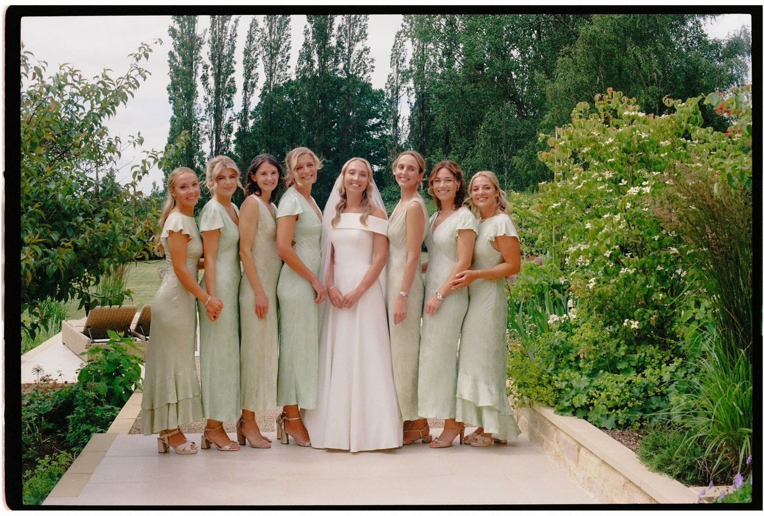 Bride in white gown with seven bridesmaids in sage green dresses posing in a lush garden setting.