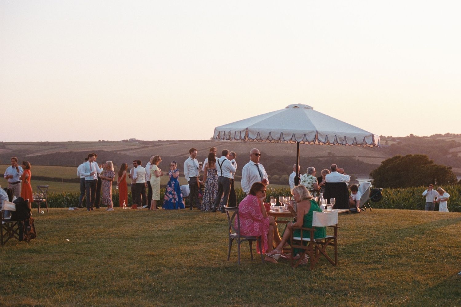 Outdoor wedding reception on green hillside at sunset with guests mingling near a white market umbrella.