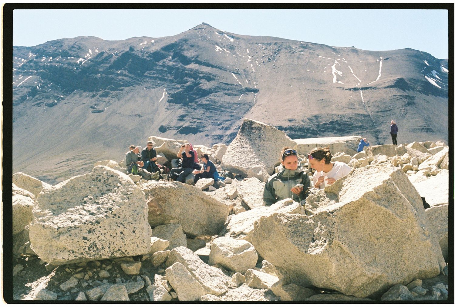 Group of hikers resting among large boulders on a rocky mountain summit with peaks in background.
