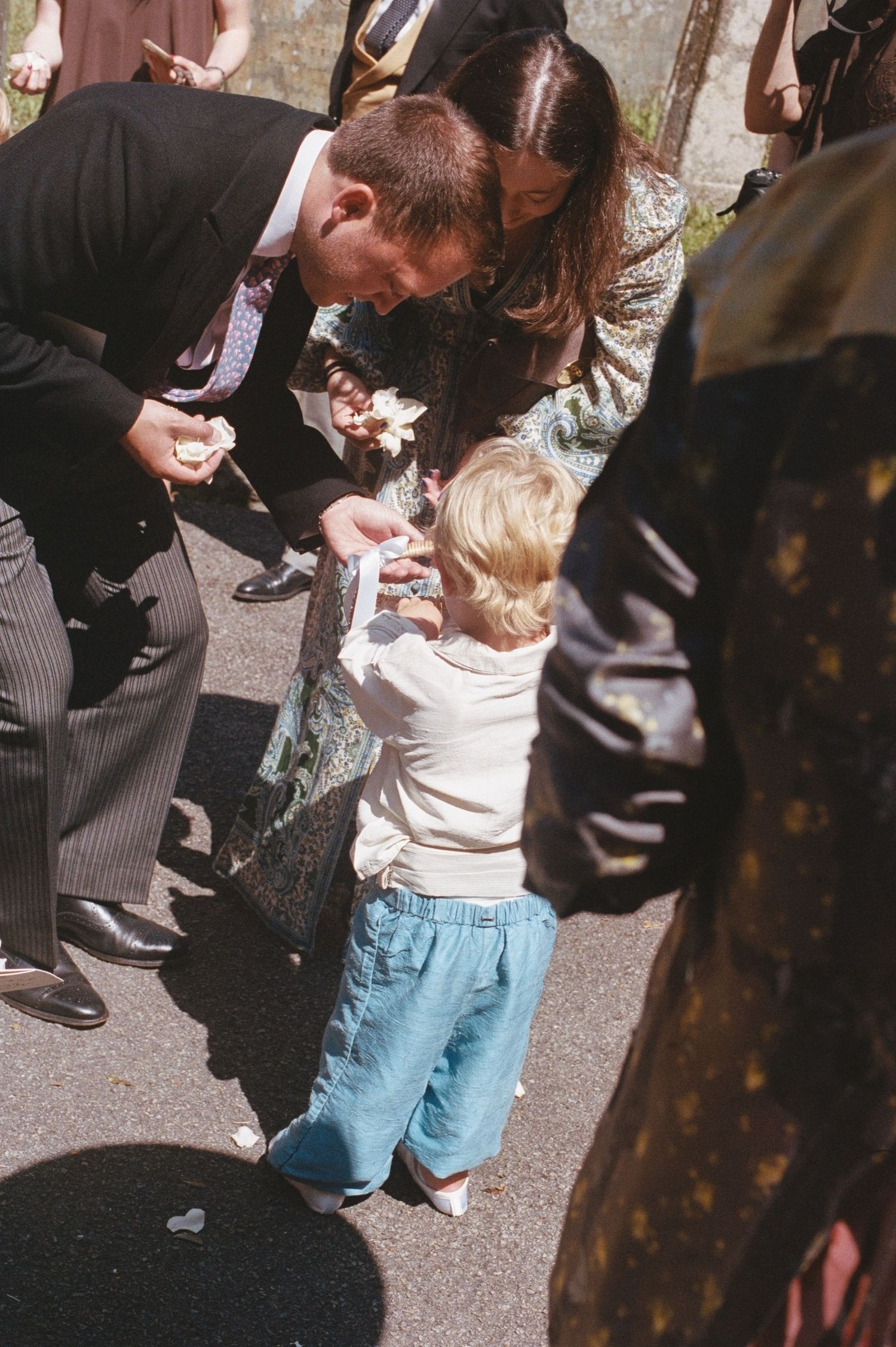 Adults in formal attire lean down to interact with a young blonde toddler outdoors at a sunny event.