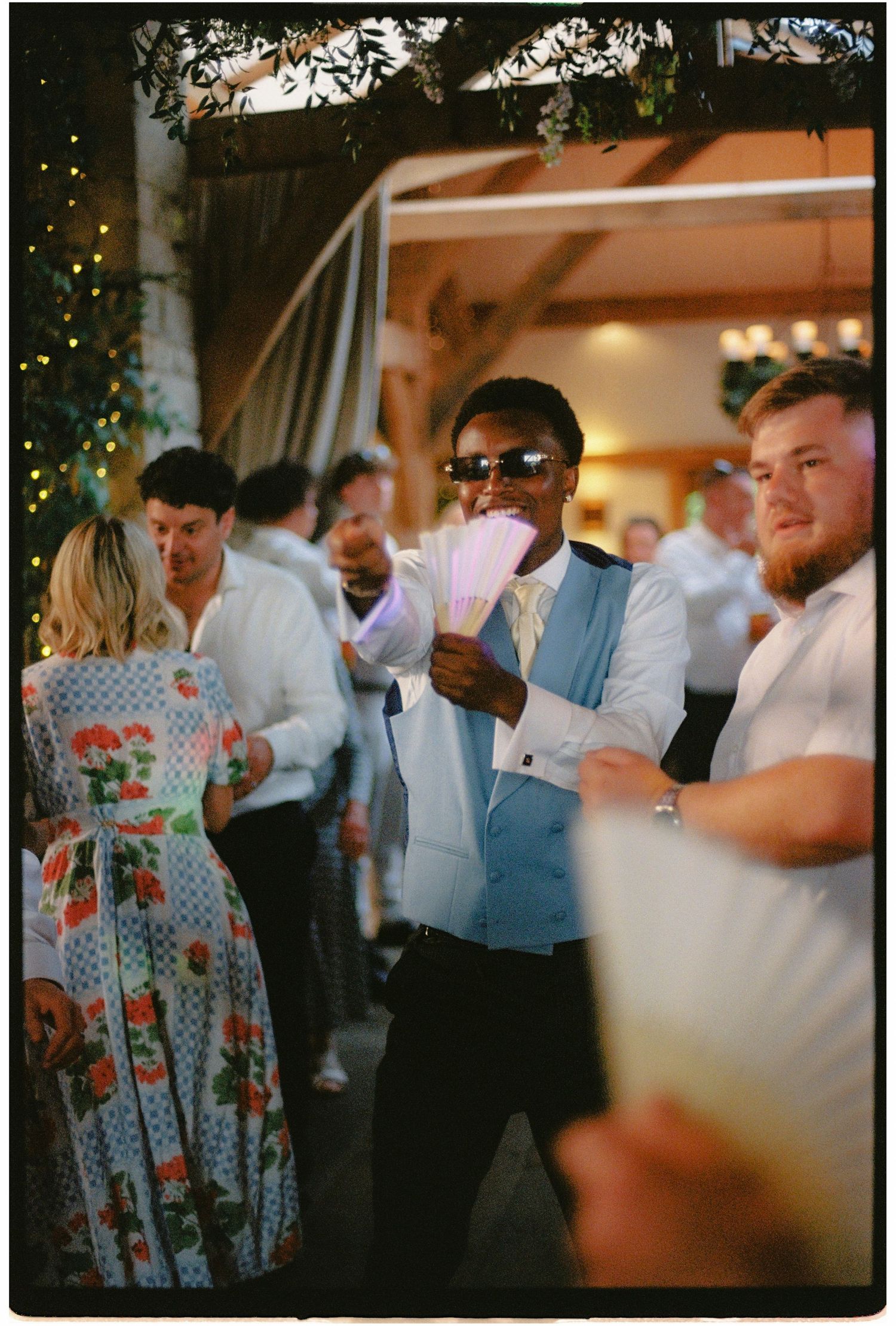 A man in a light blue vest and sunglasses holds cards at a lively wedding reception party.