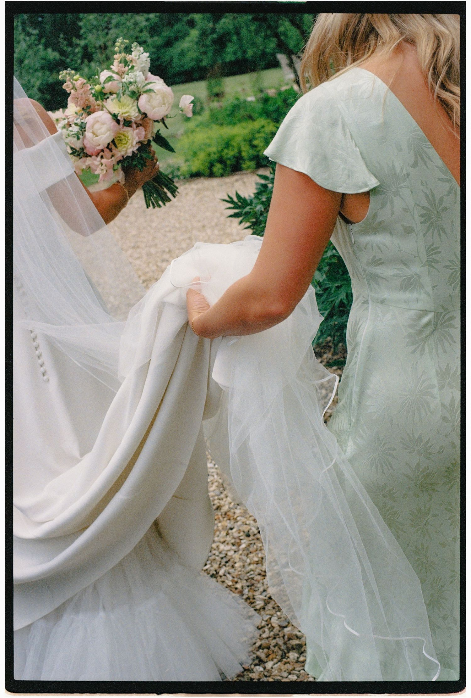 Bridesmaid in mint green dress holds bridal gown train as bride carries pink and white bouquet outdoors.